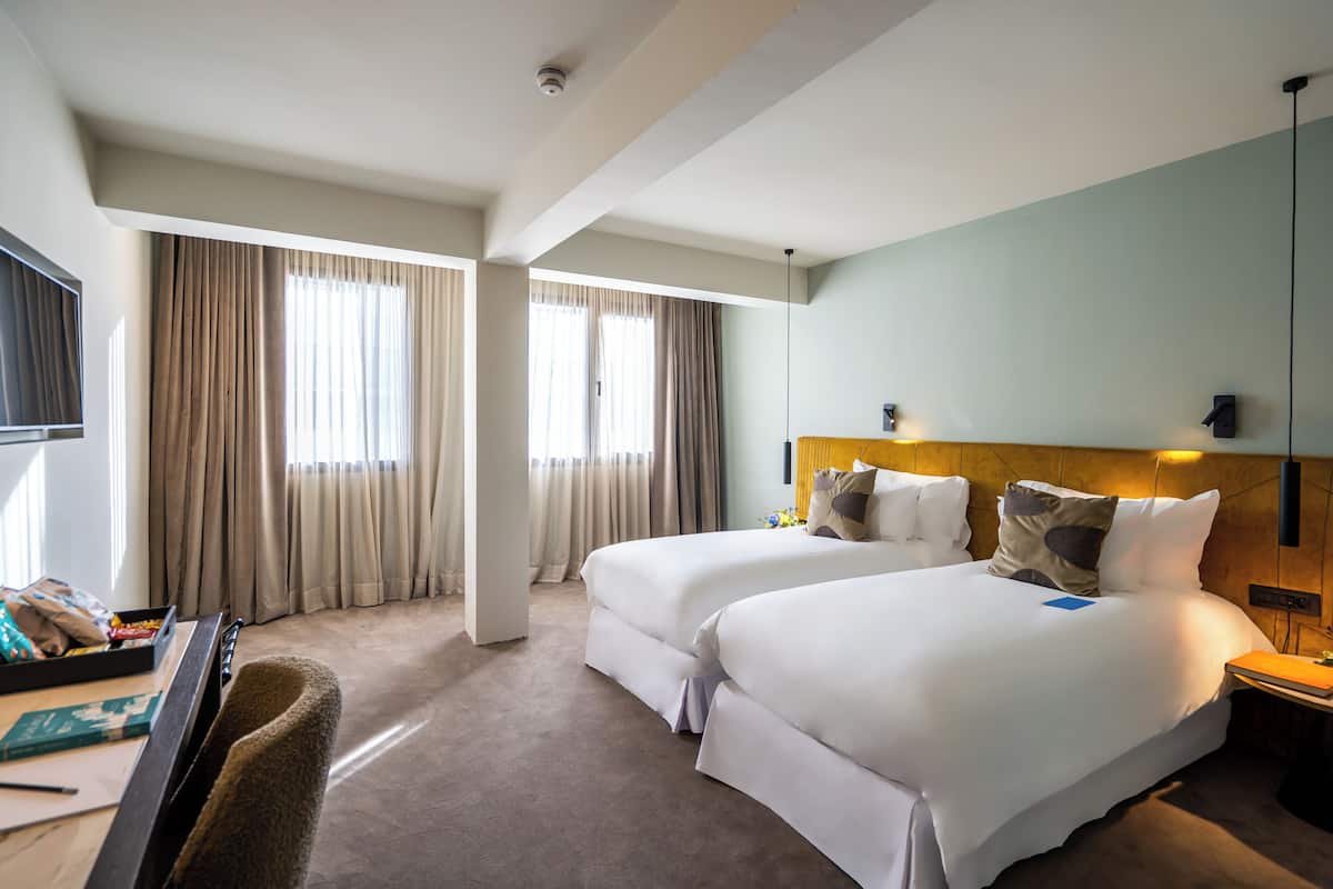 Habitación de hotel con dos camas, un escritorio con silla, un televisor de pantalla plana y un ventanal con cortinas.