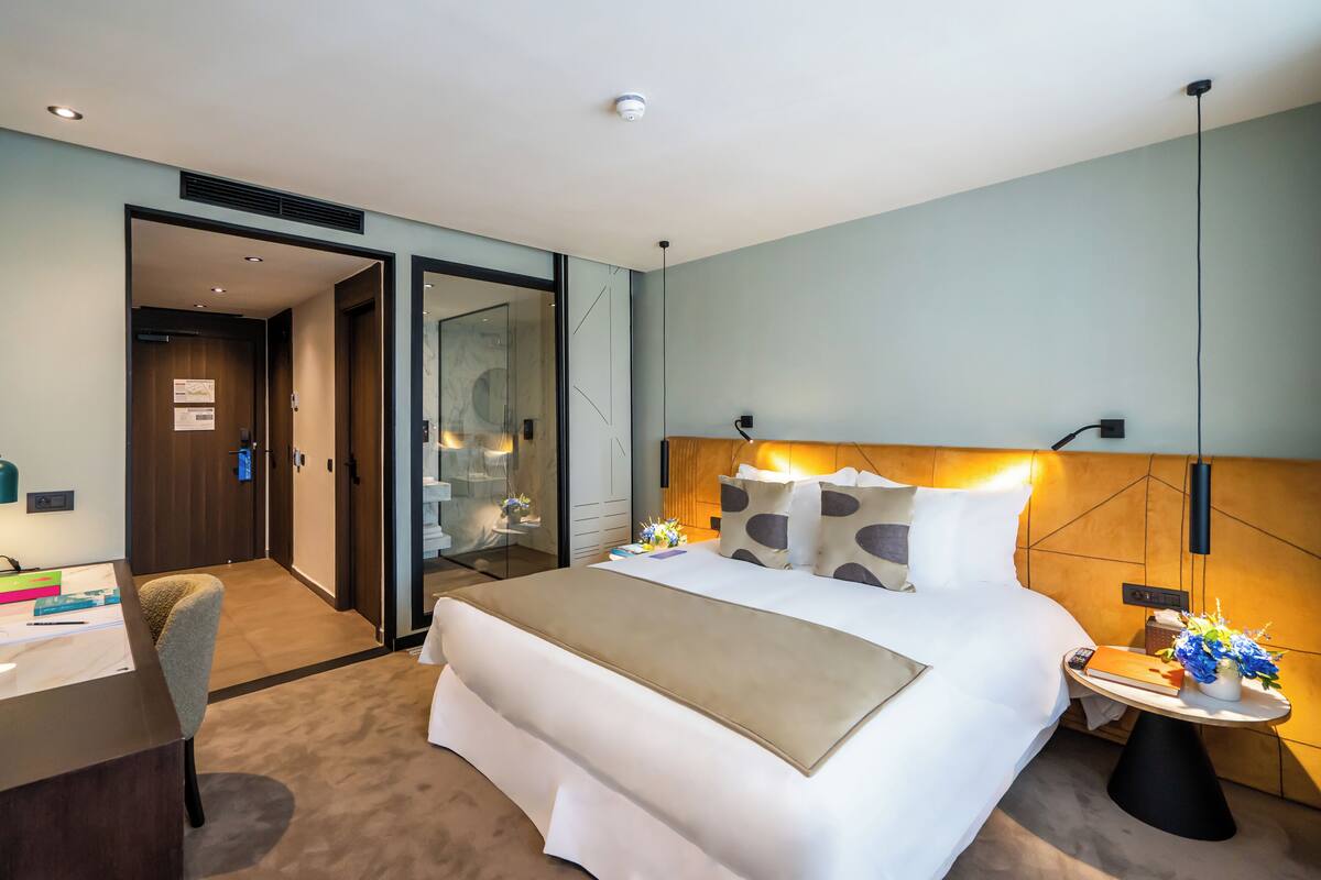 Habitación de hotel con cama, mesita de noche, escritorio, silla y vista al pasillo.