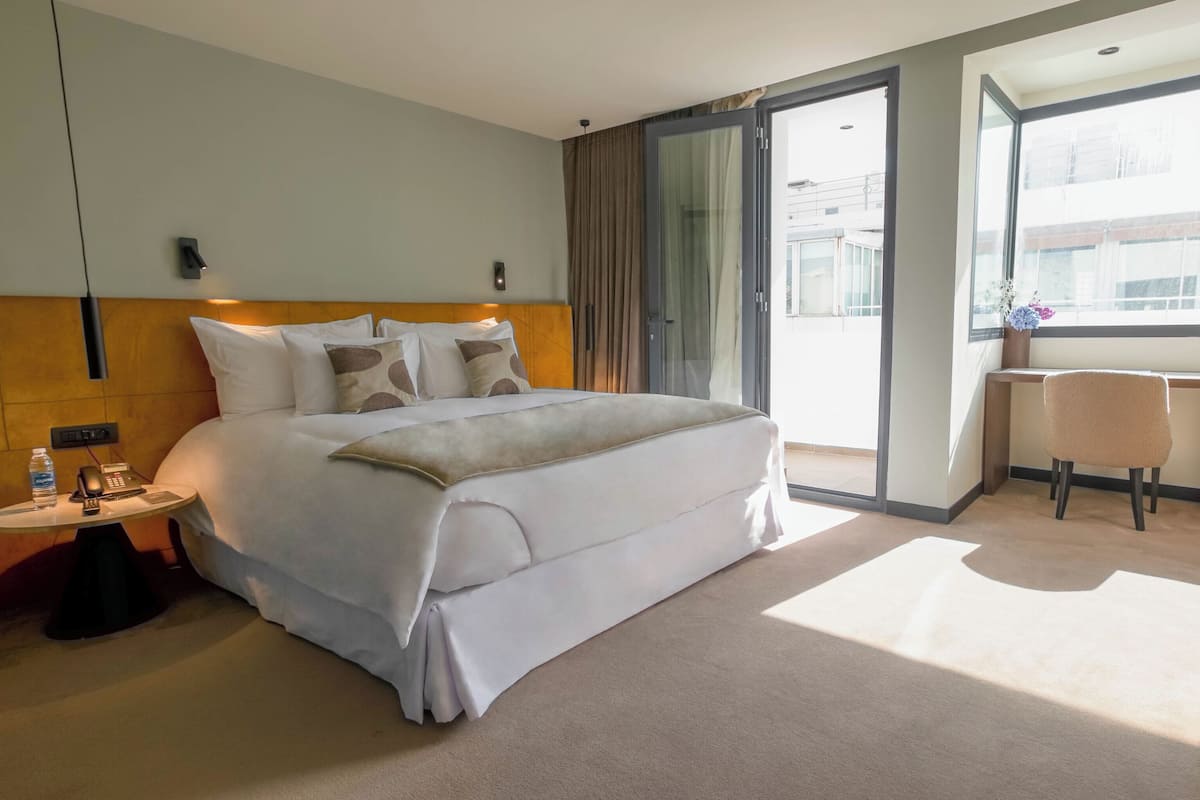 Habitación de hotel con cama, mesita de noche con una botella y teléfono, escritorio con silla y un ventanal grande.
