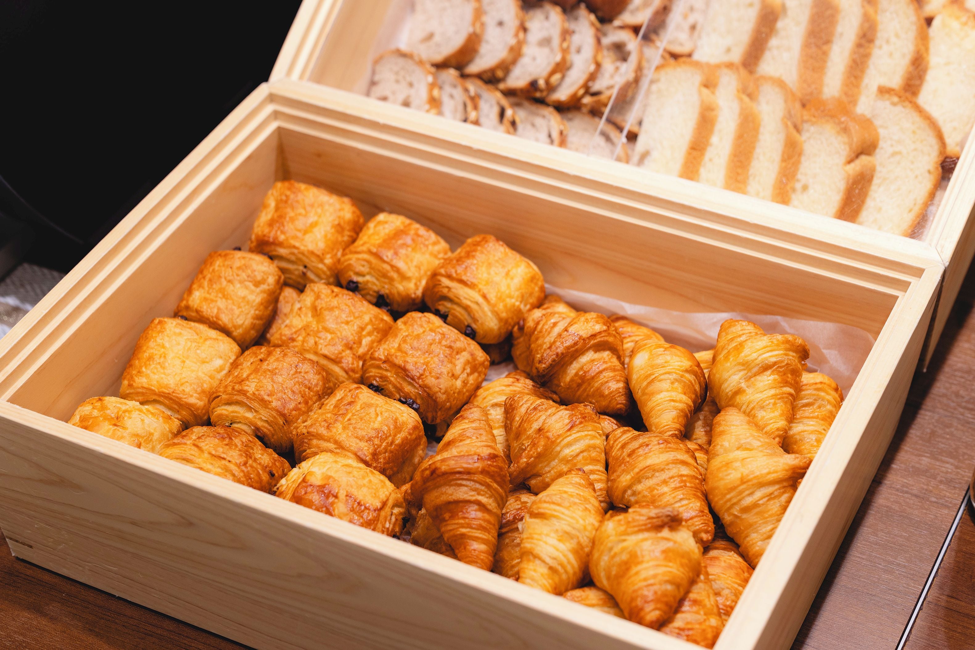 Een houten doos gevuld met gebakjes, waaronder croissants en sneetjes brood.