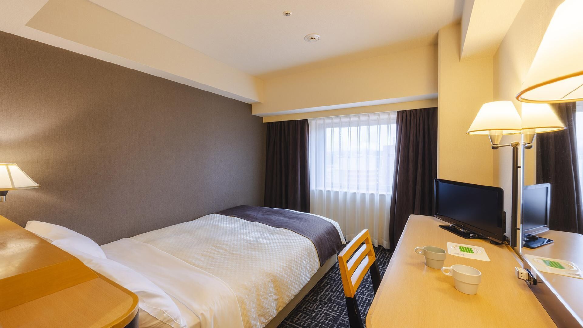 A hotel room with a bed, a desk with a computer, a chair, and a lamp.