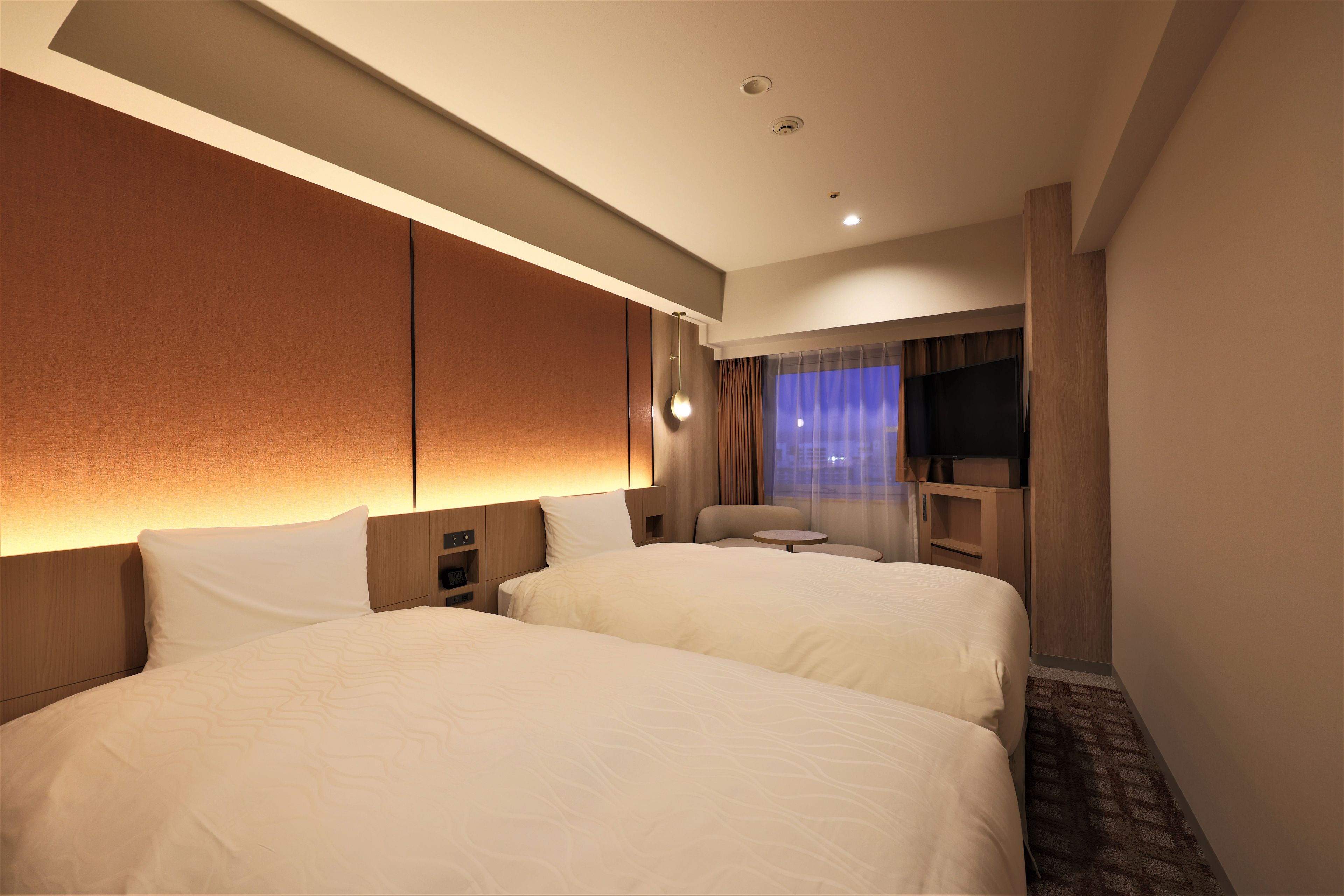 A hotel room with two beds, a desk, a chair, a TV, and a window with curtains.