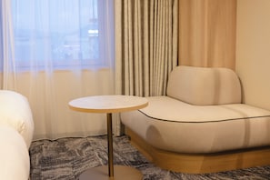 A hotel room with a sofa, a small round table, and a view of the city through a window with curtains. - Canal City Fukuoka Washington Hotel (Fukuoka)