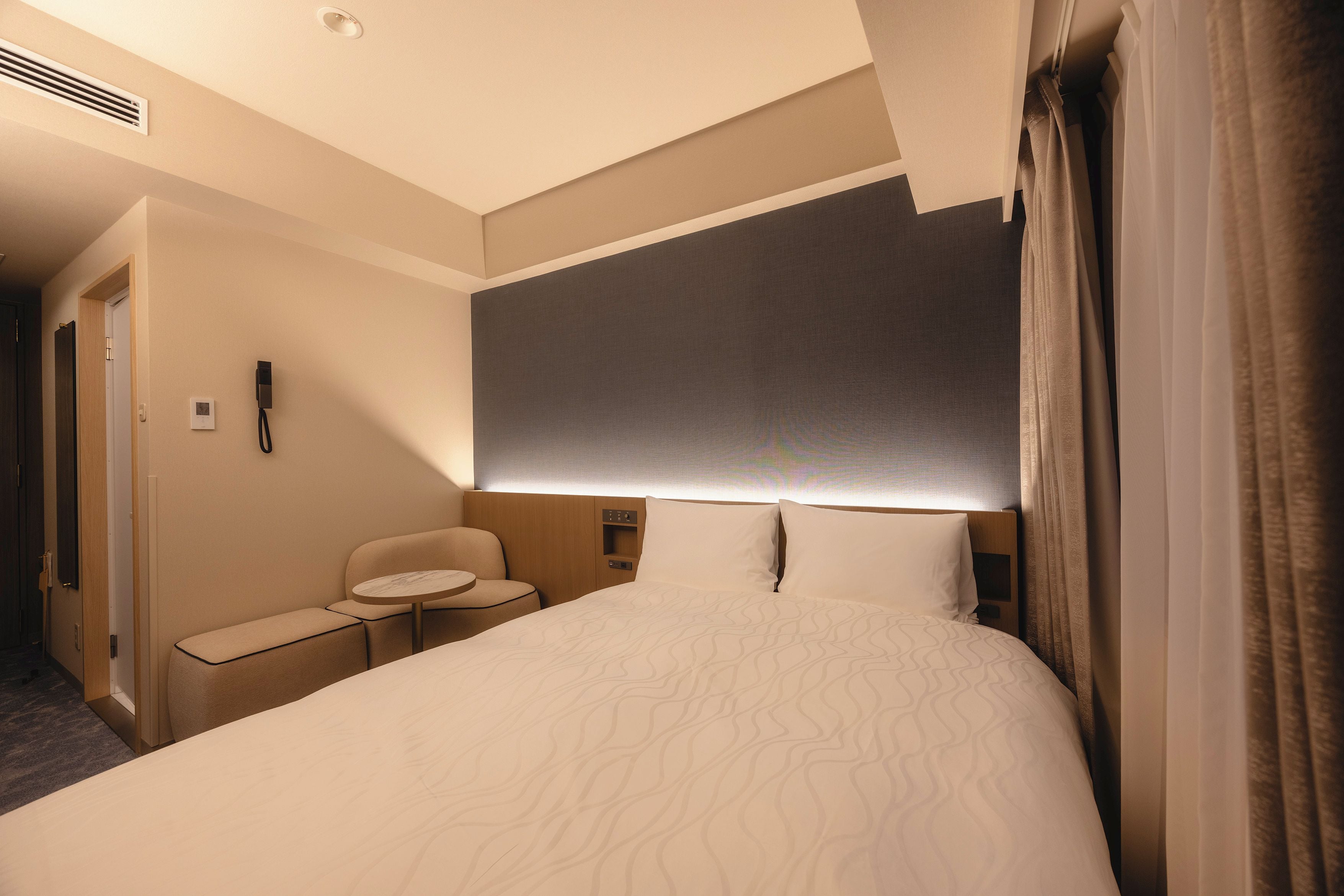 A hotel room with a large bed, a bedside table, a chair, a small table, and a phone on the wall.
