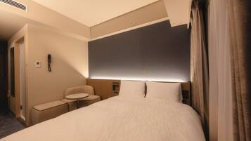 A hotel room with a large bed, a bedside table, a chair, a small table, and a phone on the wall.