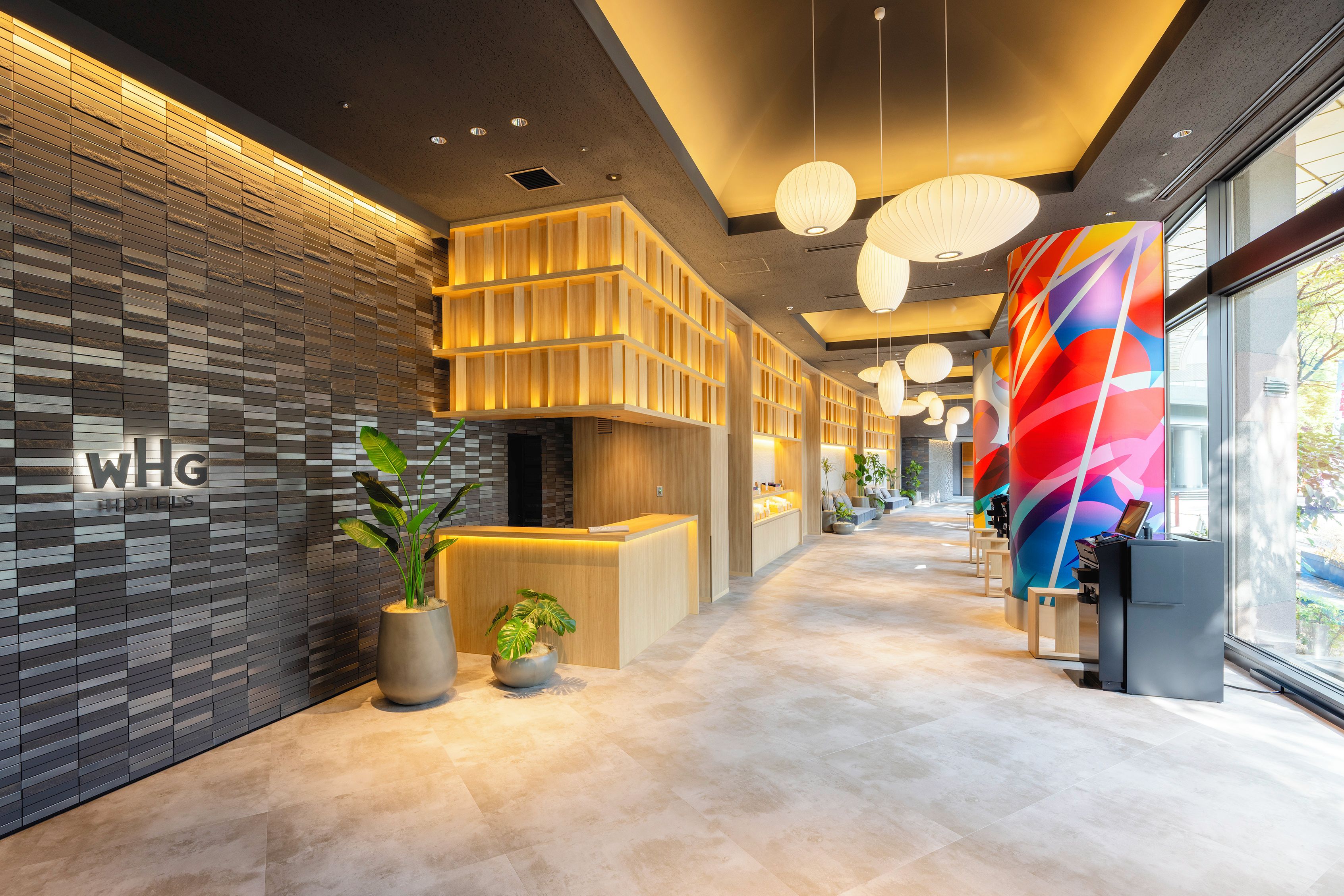 A modern hotel reception area with a reception desk, potted plants, and a colorful wall mural.