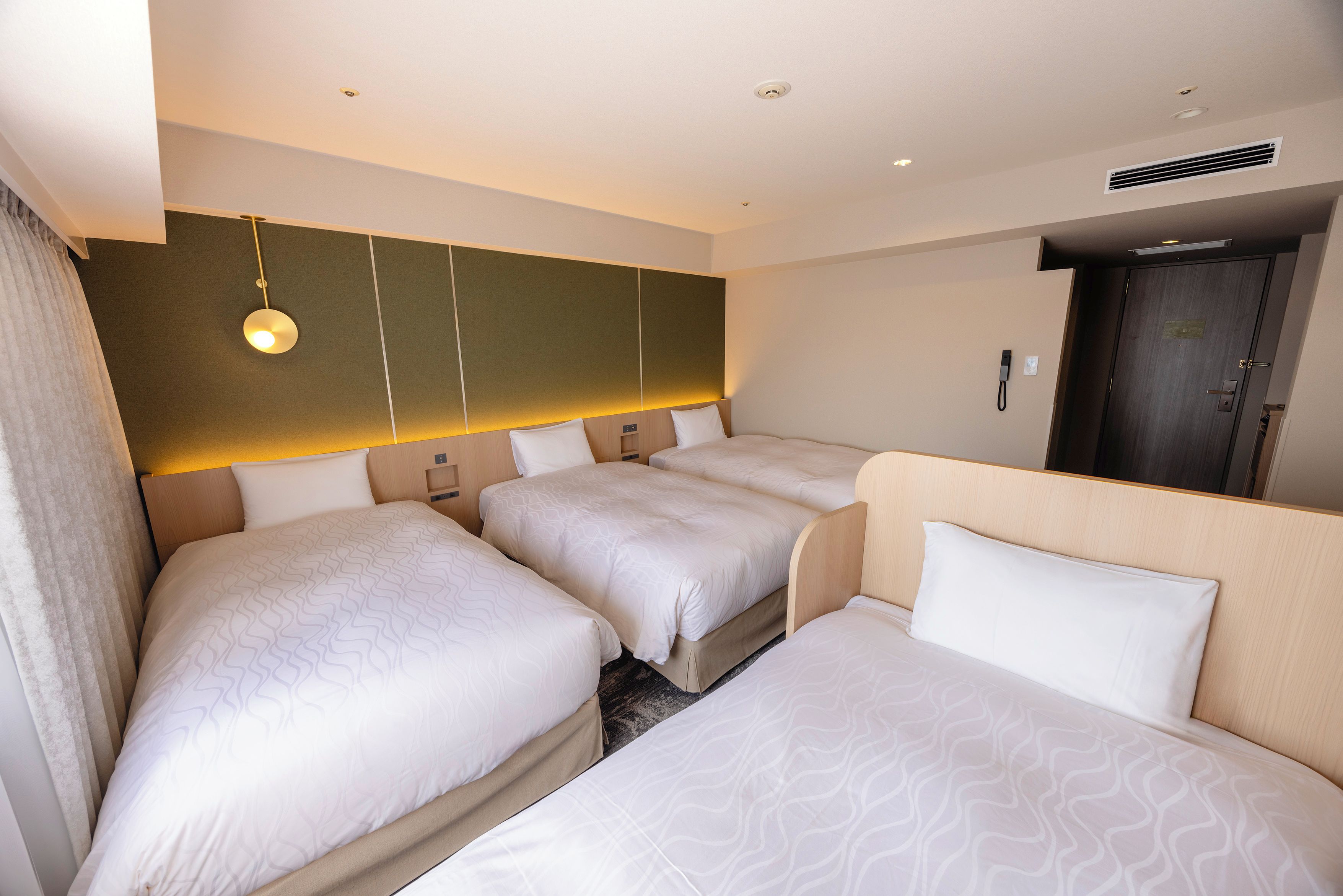Habitación de hotel con tres camas, una lámpara fijada a la pared, una puerta con cerradura y una rejilla en el techo.
