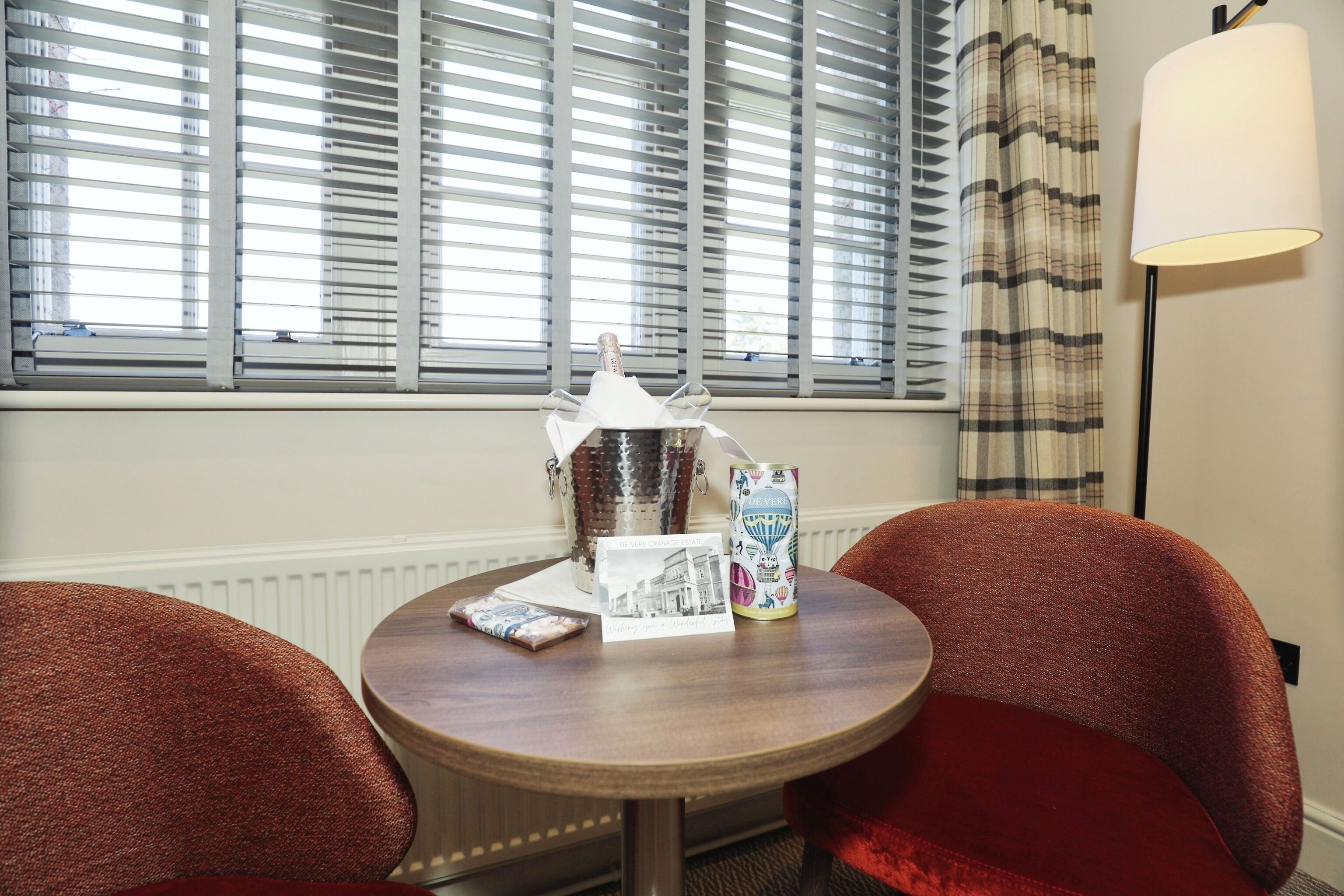 A hotel room with a round wooden table, two red upholstered chairs, a window with blinds, a lamp, and a minibar.