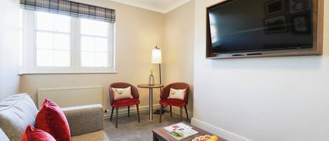 Un salon comprenant un canapé, deux fauteuils, une table basse avec des fruits, une lampe et un téléviseur fixé au mur.