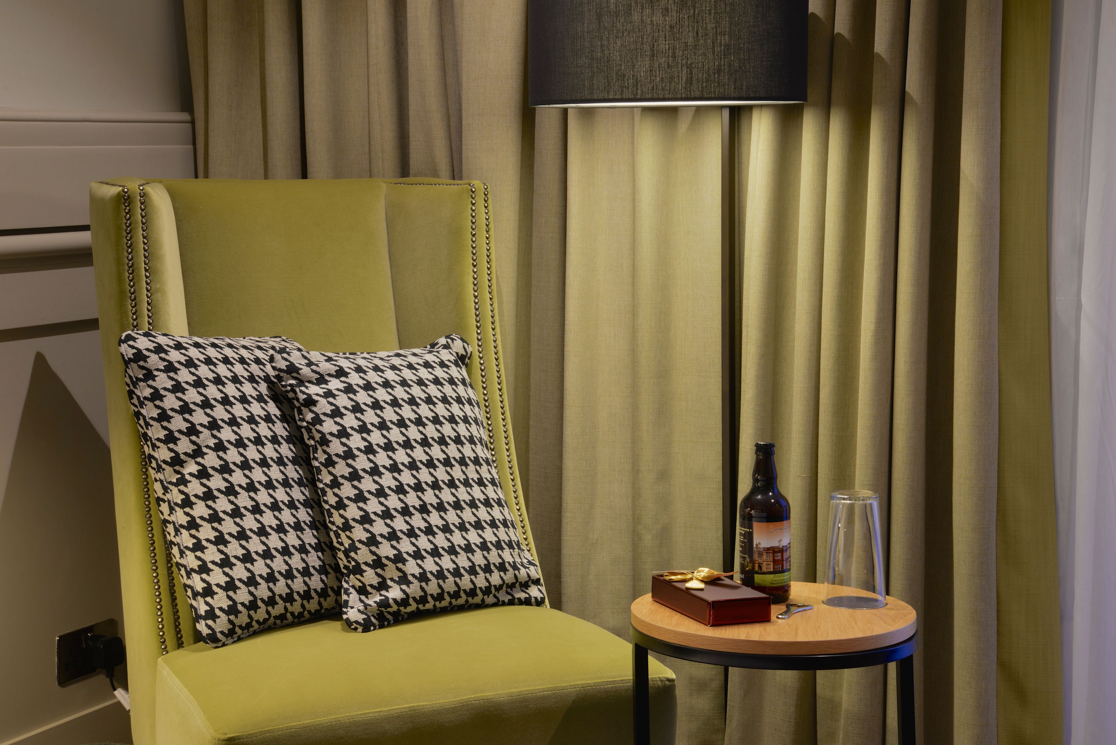 A green armchair with houndstooth pillows, a small round table with a bottle, glasses, and a box.