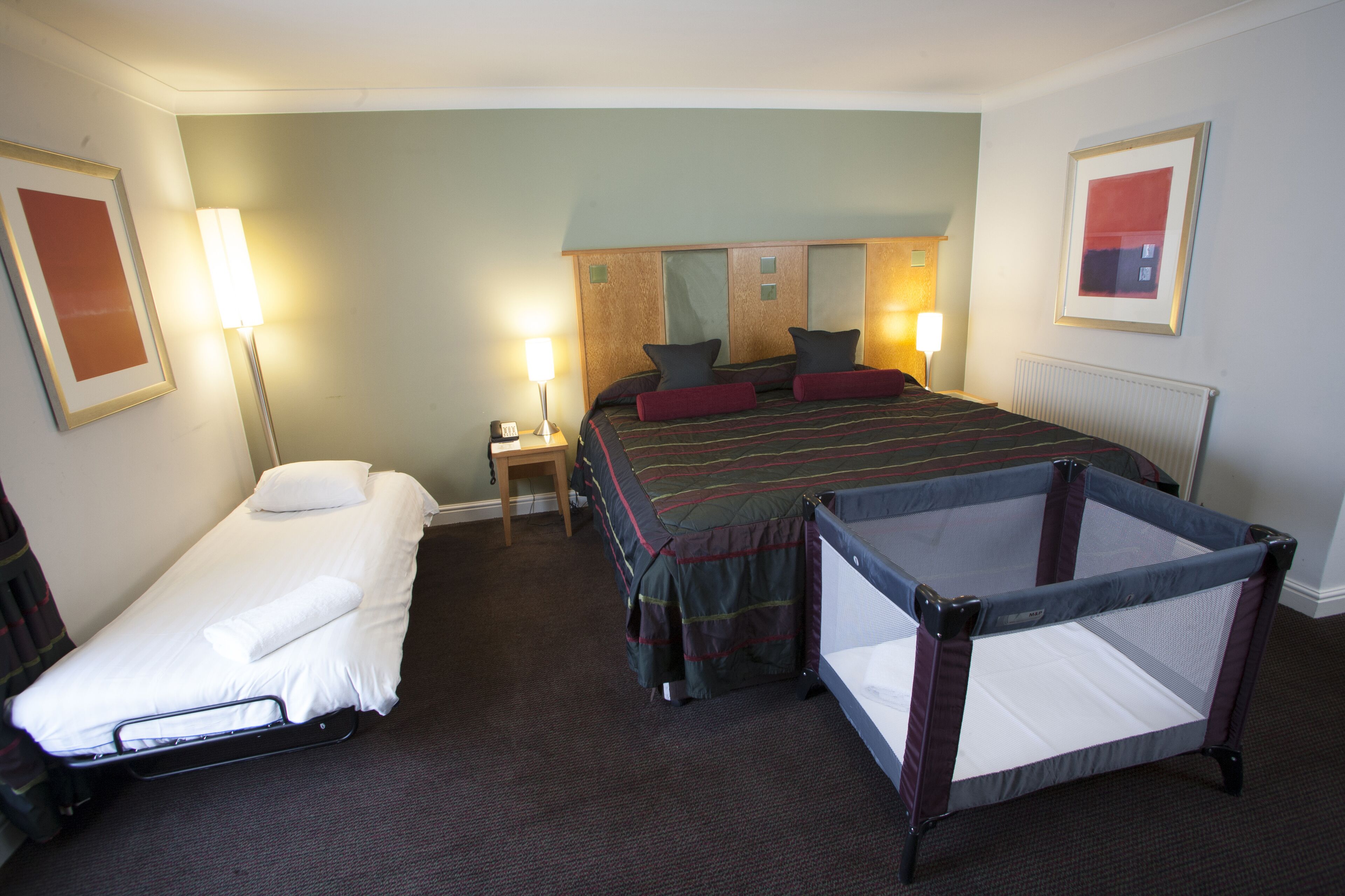 Une chambre d’hôtel avec un lit simple, un canapé-lit, un berceau et une table de chevet avec une lampe.