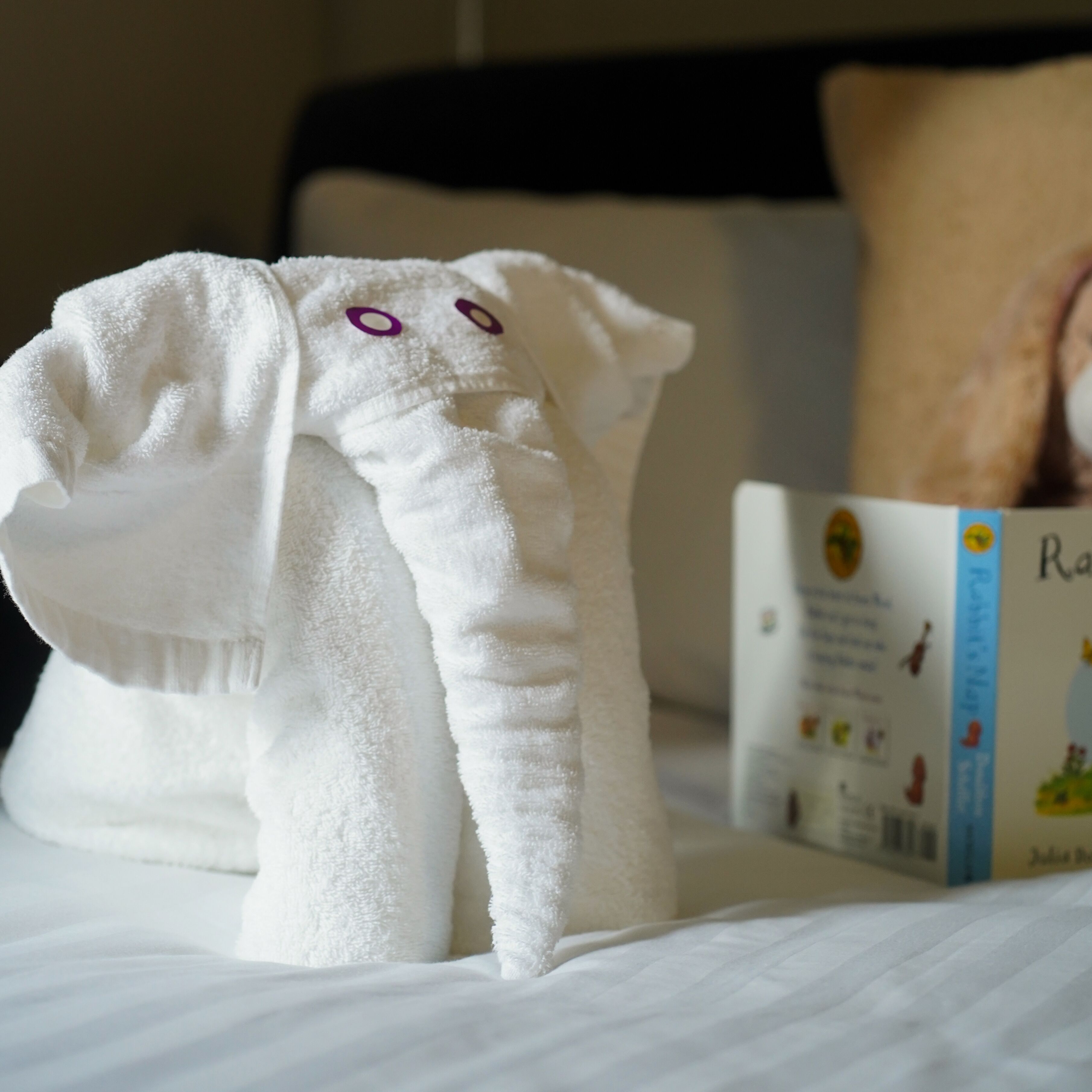 A neatly made towel shaped like an elephant on a bed with a pillow and a book.