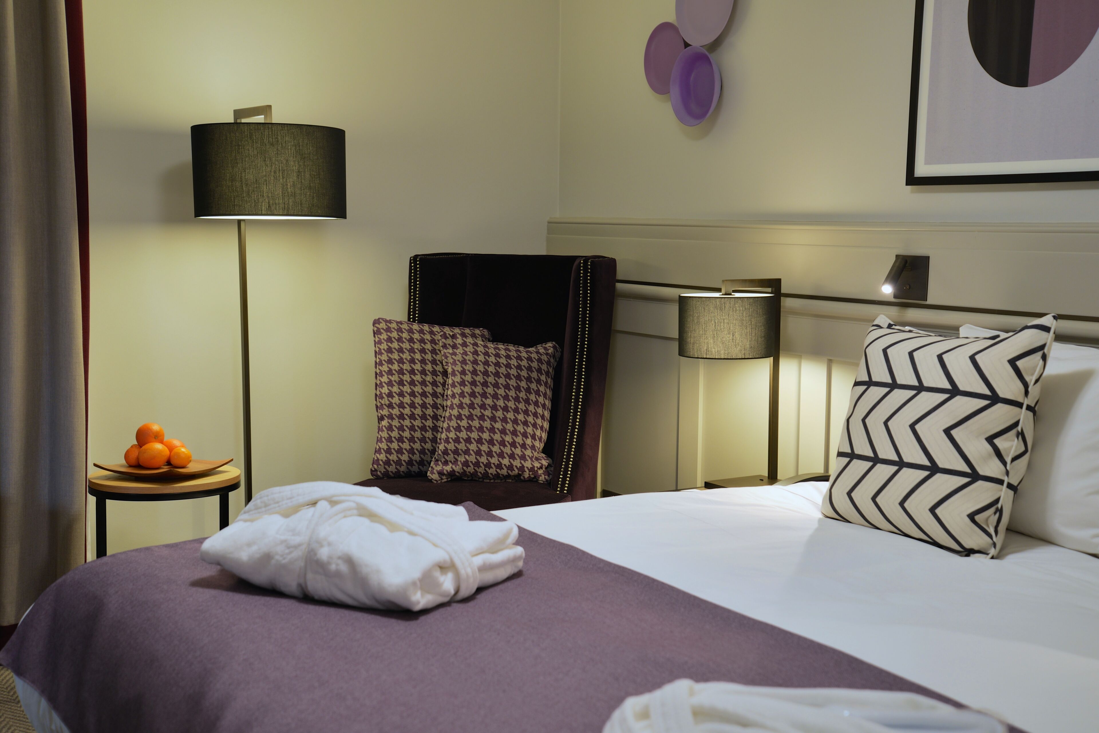 A hotel room with a neatly made bed, a chair with a patterned cushion, a small table with fruit, and a wall-mounted lamp.