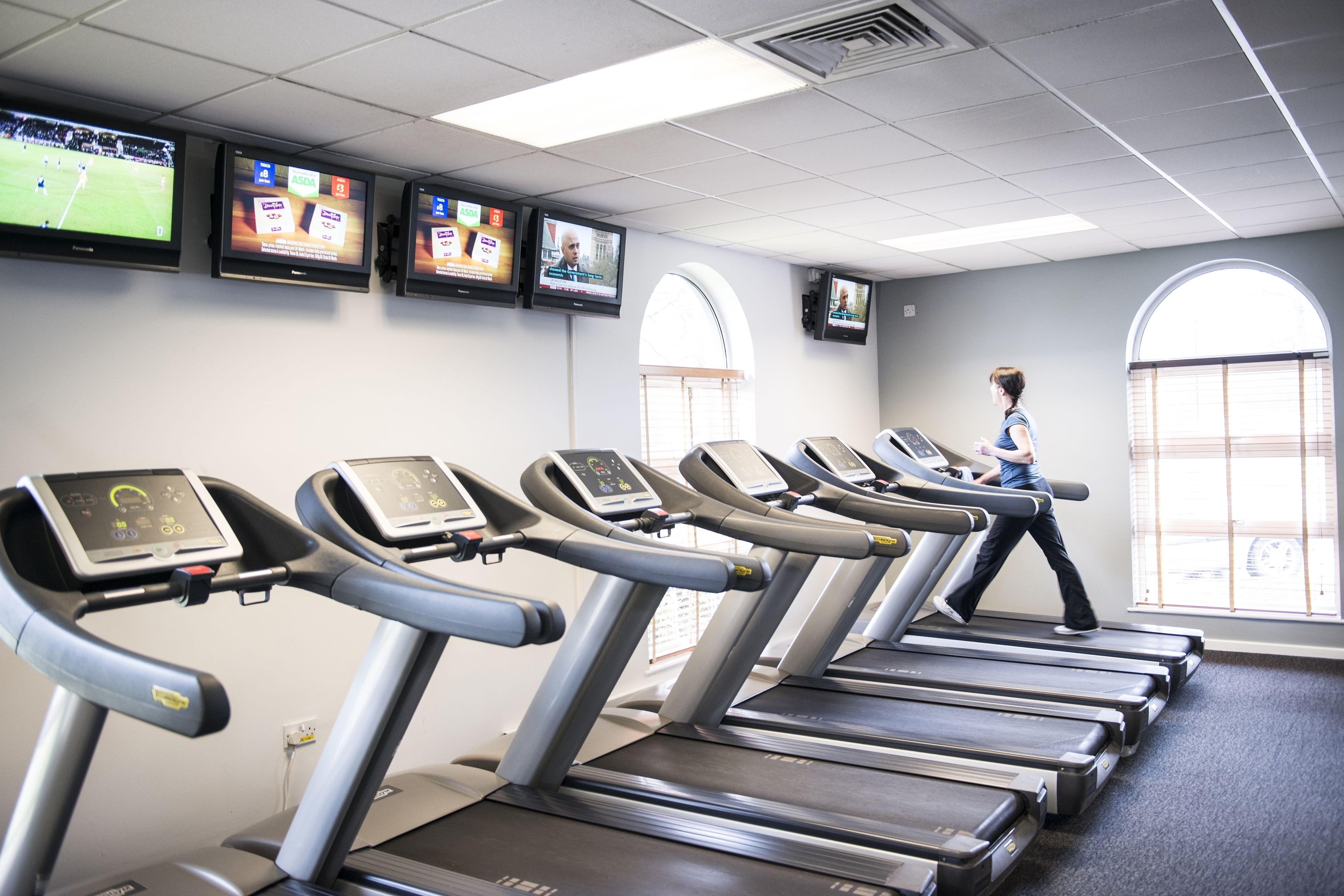 Une salle de sport équipée de plusieurs tapis de course, où une femme marche sur l’un d’eux, et plusieurs téléviseurs fixés au mur diffusent différents programmes.