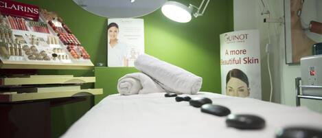 Um quarto de spa com uma mesa de massagem, uma exposição de produtos de cuidado da pele Clarins e um cartaz divulgando tratamentos de pele Guinot.
