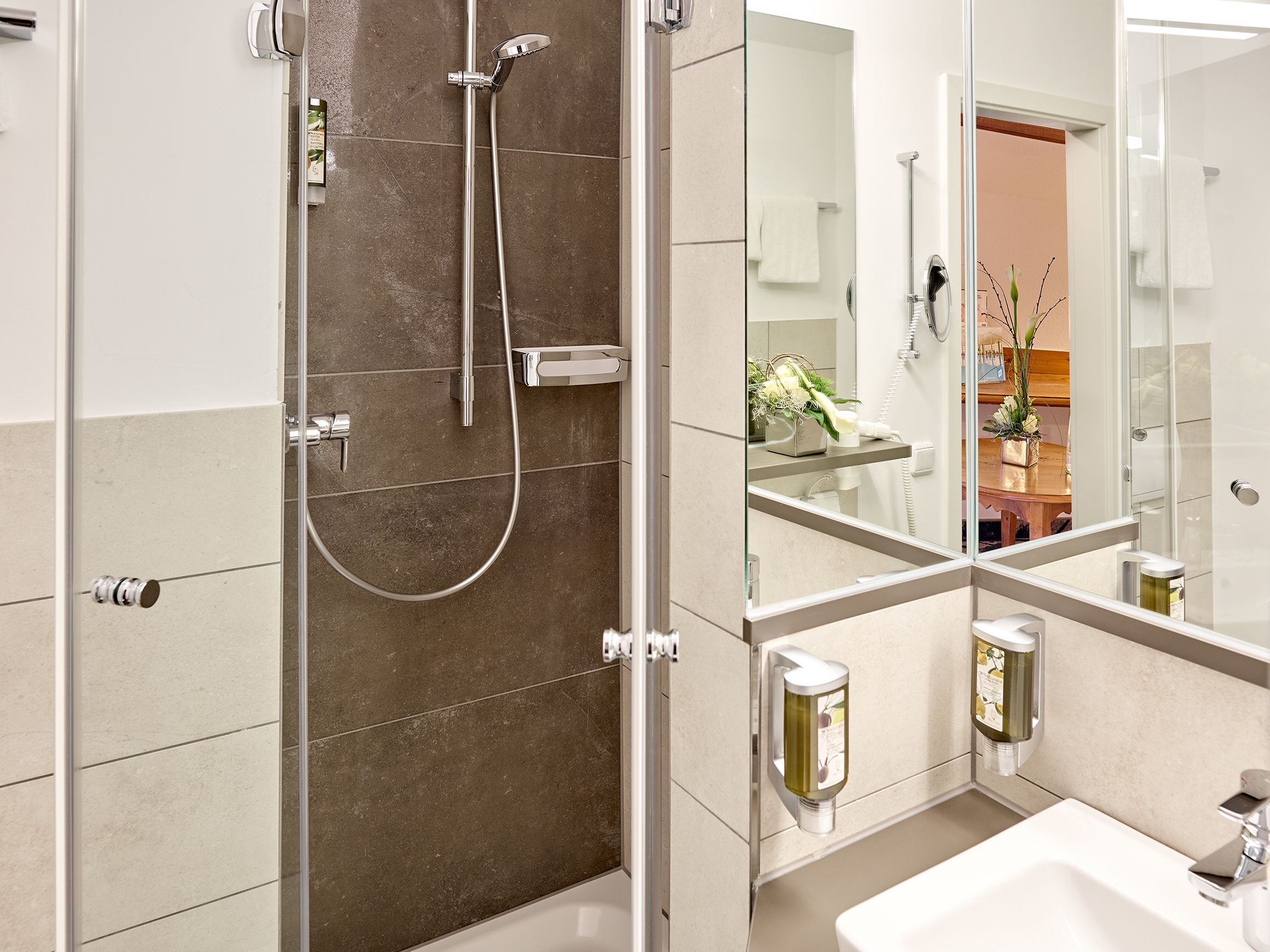 A modern bathroom with a glass shower enclosure, a white sink, and a mirror reflecting a plant and a wooden door.