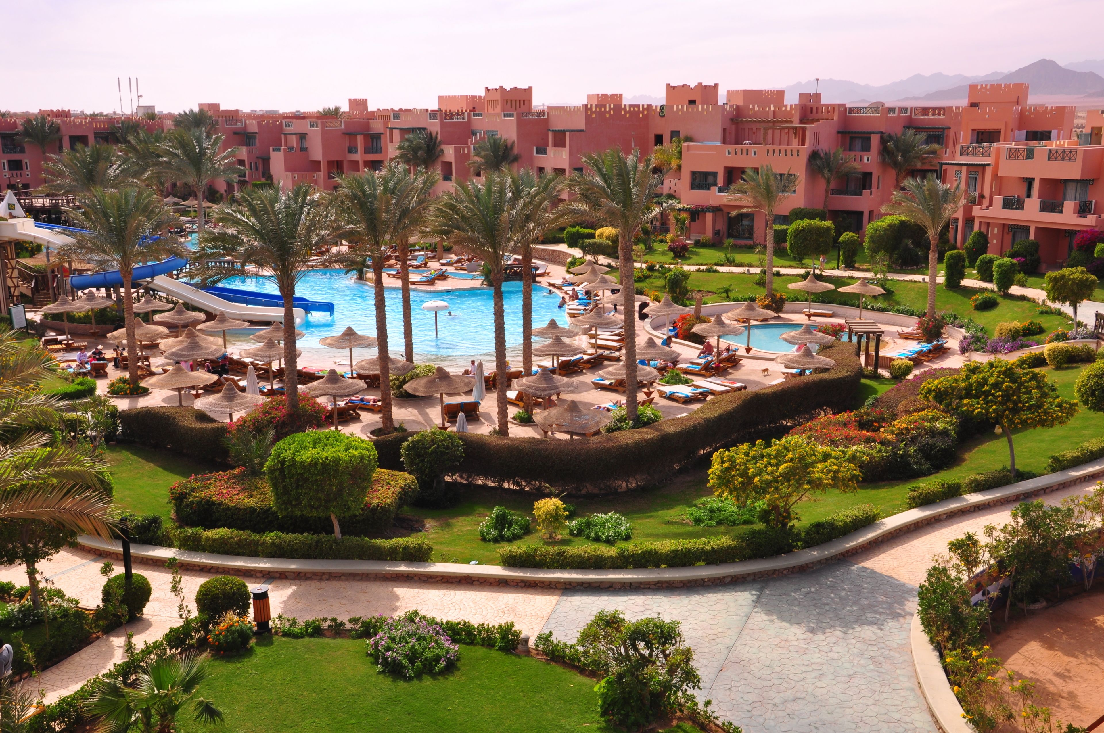 Un complejo turístico con piscina central, palmeras y un jardín ajardinado.