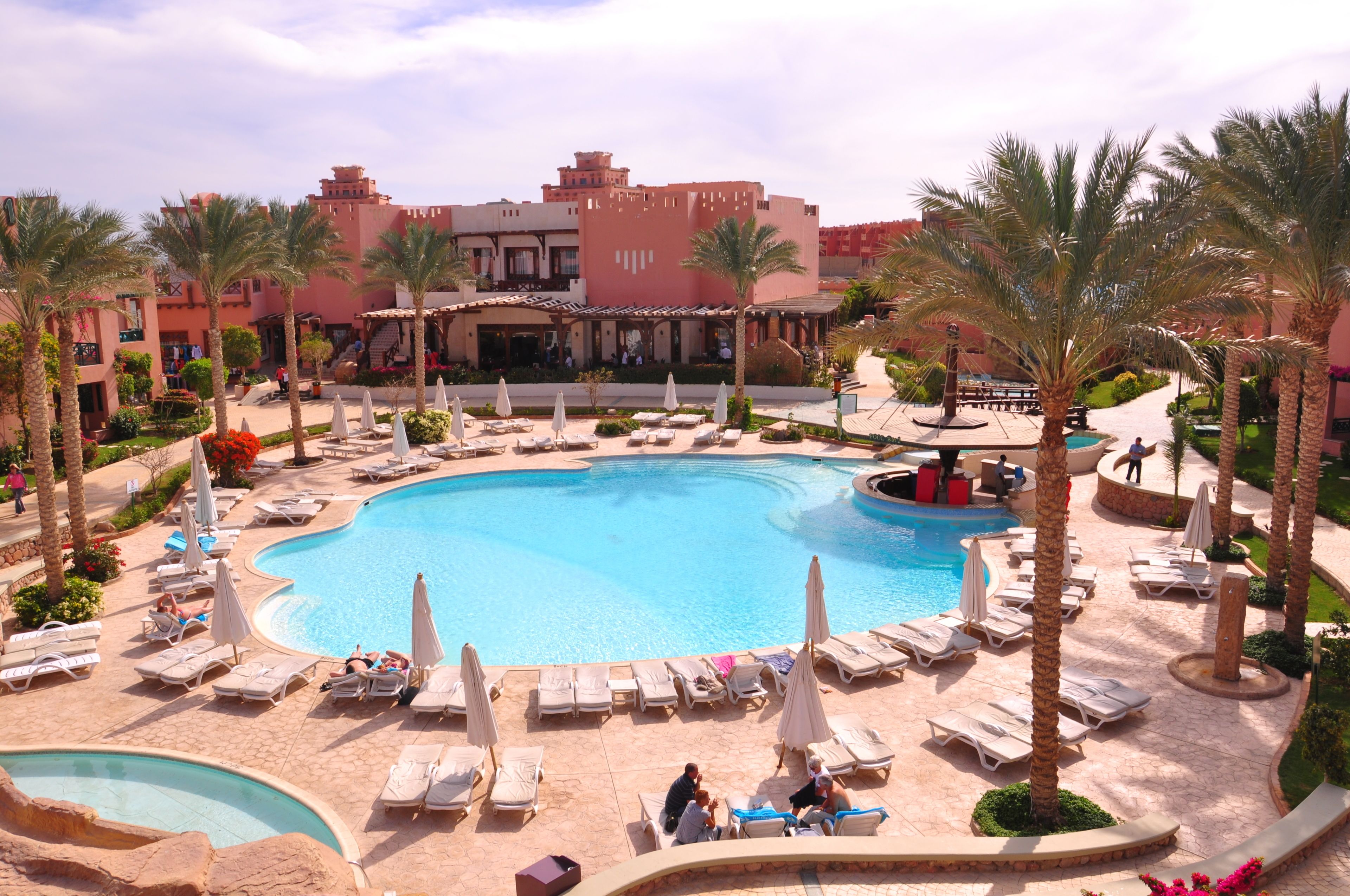 Zona de piscina en un resort con sillones de descanso y sombrillas, rodeada de palmeras y un edificio grande al fondo.