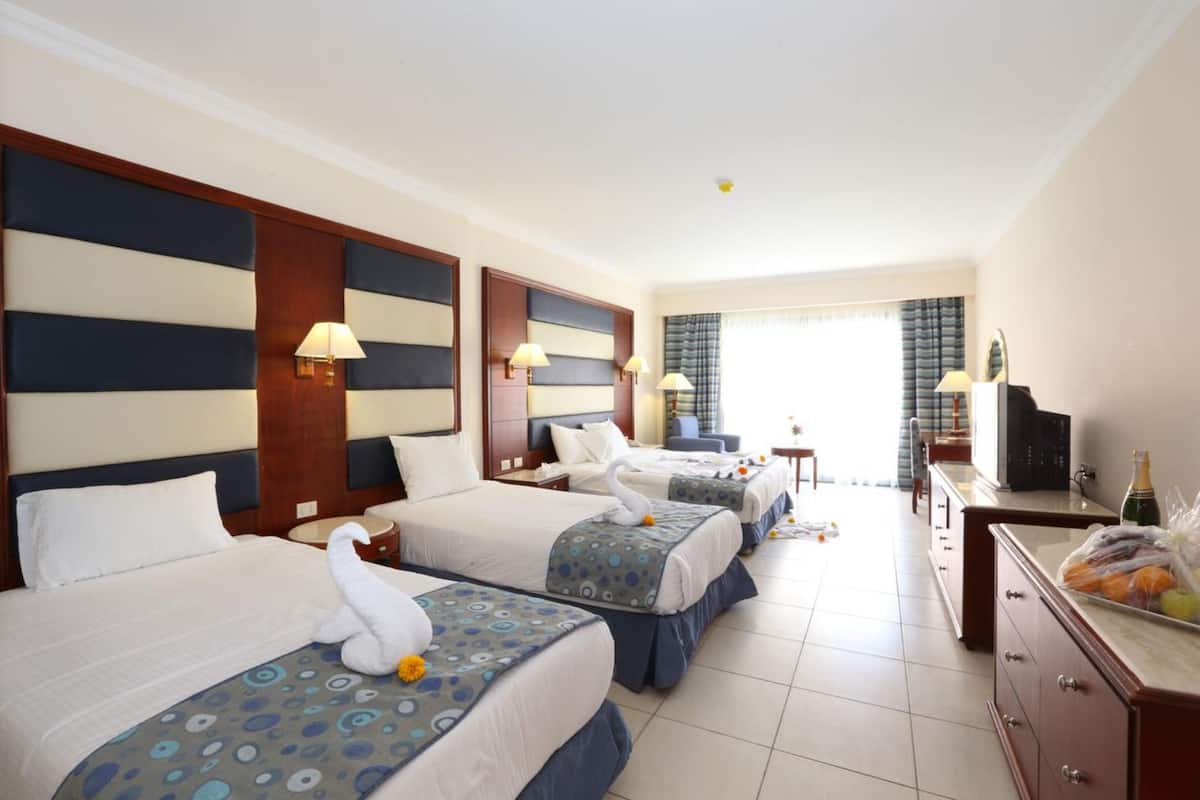 A hotel room with three beds, each with a swan-shaped pillow and a blue patterned bedspread. There is a minibar and a television on a wooden cabinet.