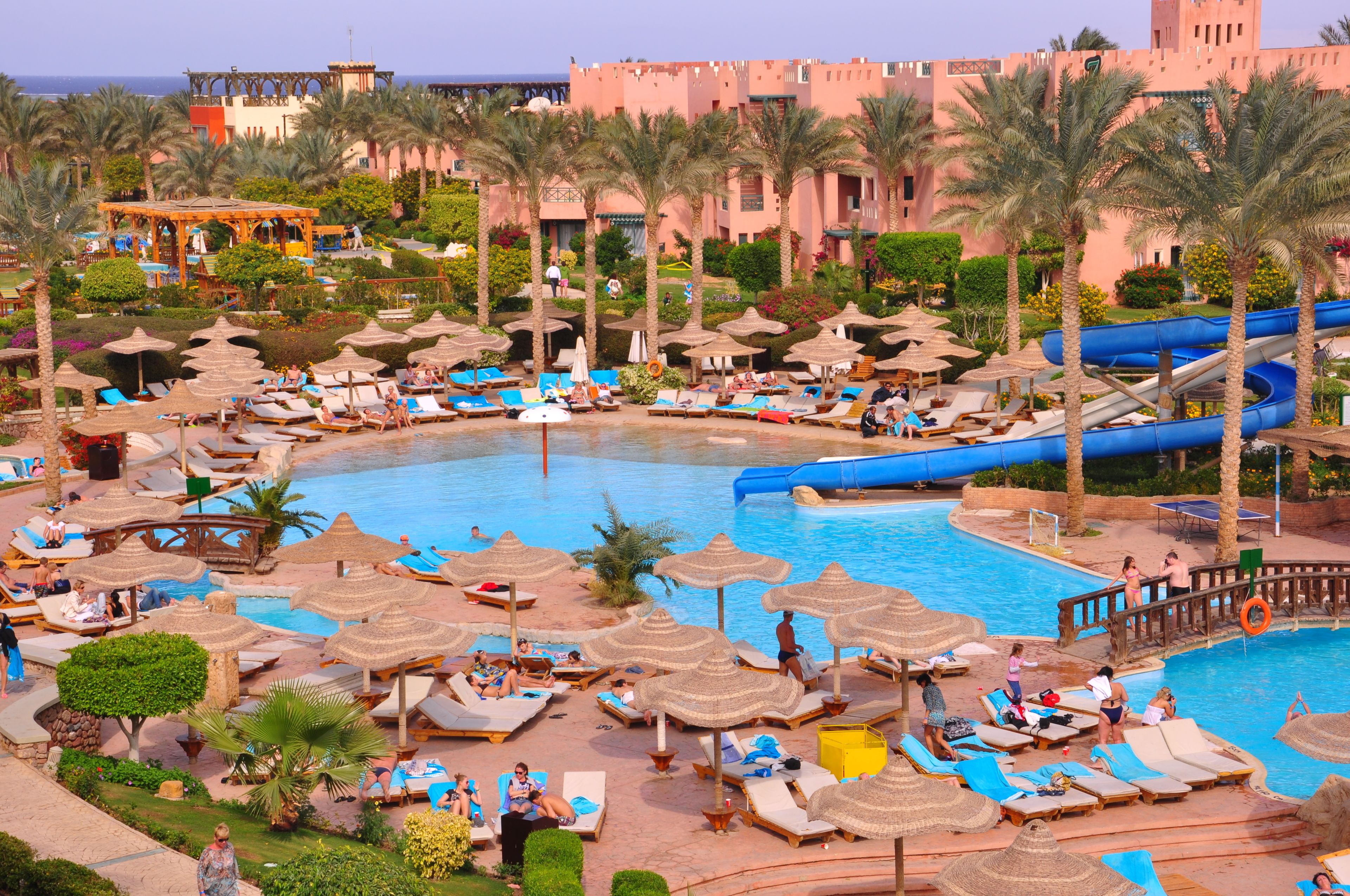 A resort pool area with lounge chairs and umbrellas, a water slide, and palm trees.