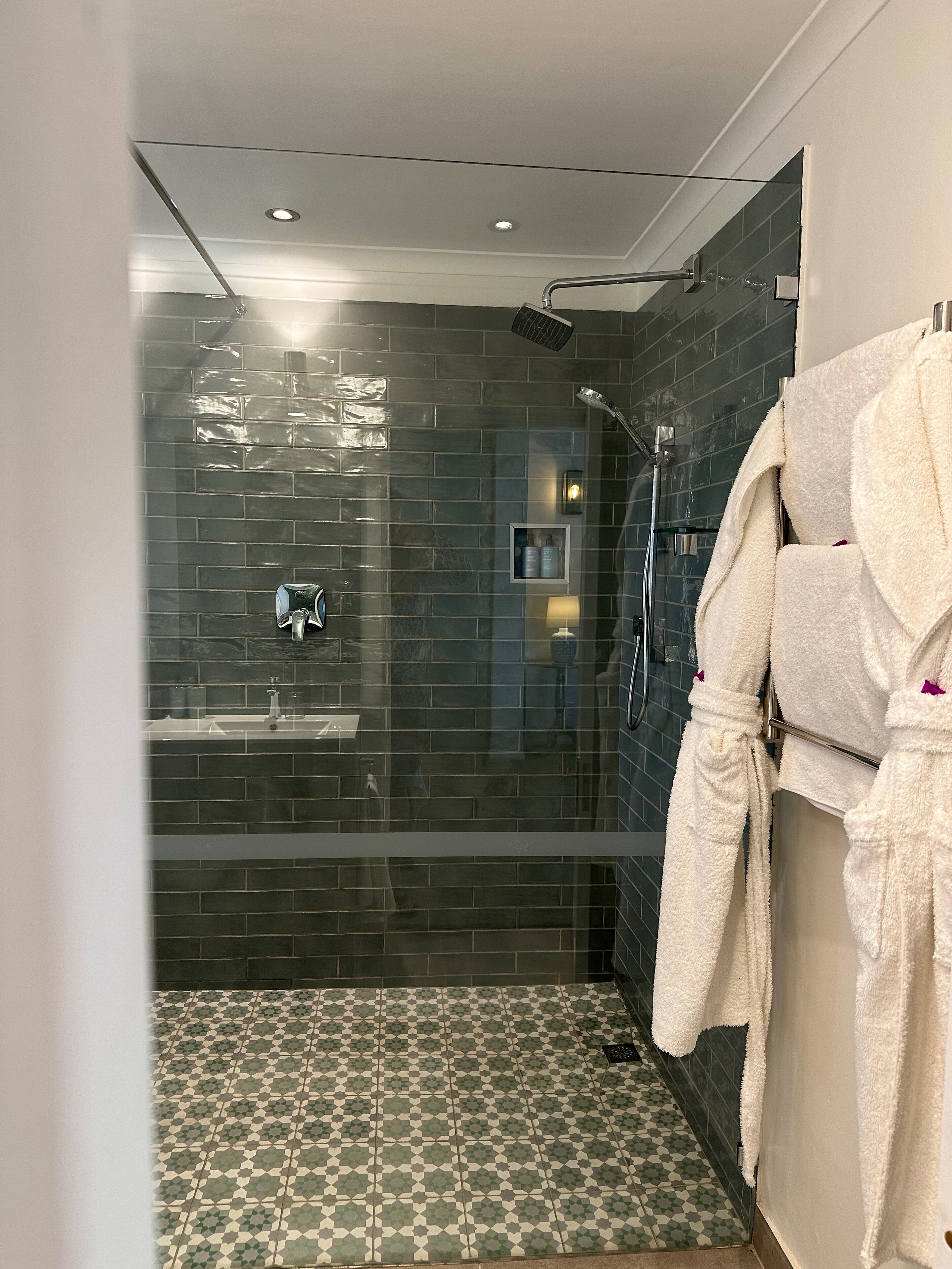 A modern bathroom with dark tiled walls, a patterned floor, a glass shower enclosure, and a towel rack with a robe.