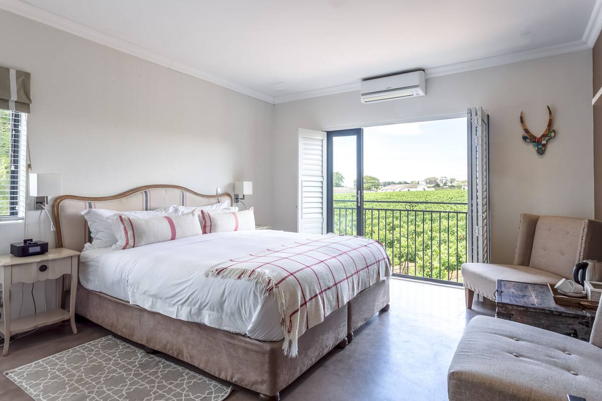 A bedroom with a large bed, a nightstand, a chair, and a balcony with a view of greenery.