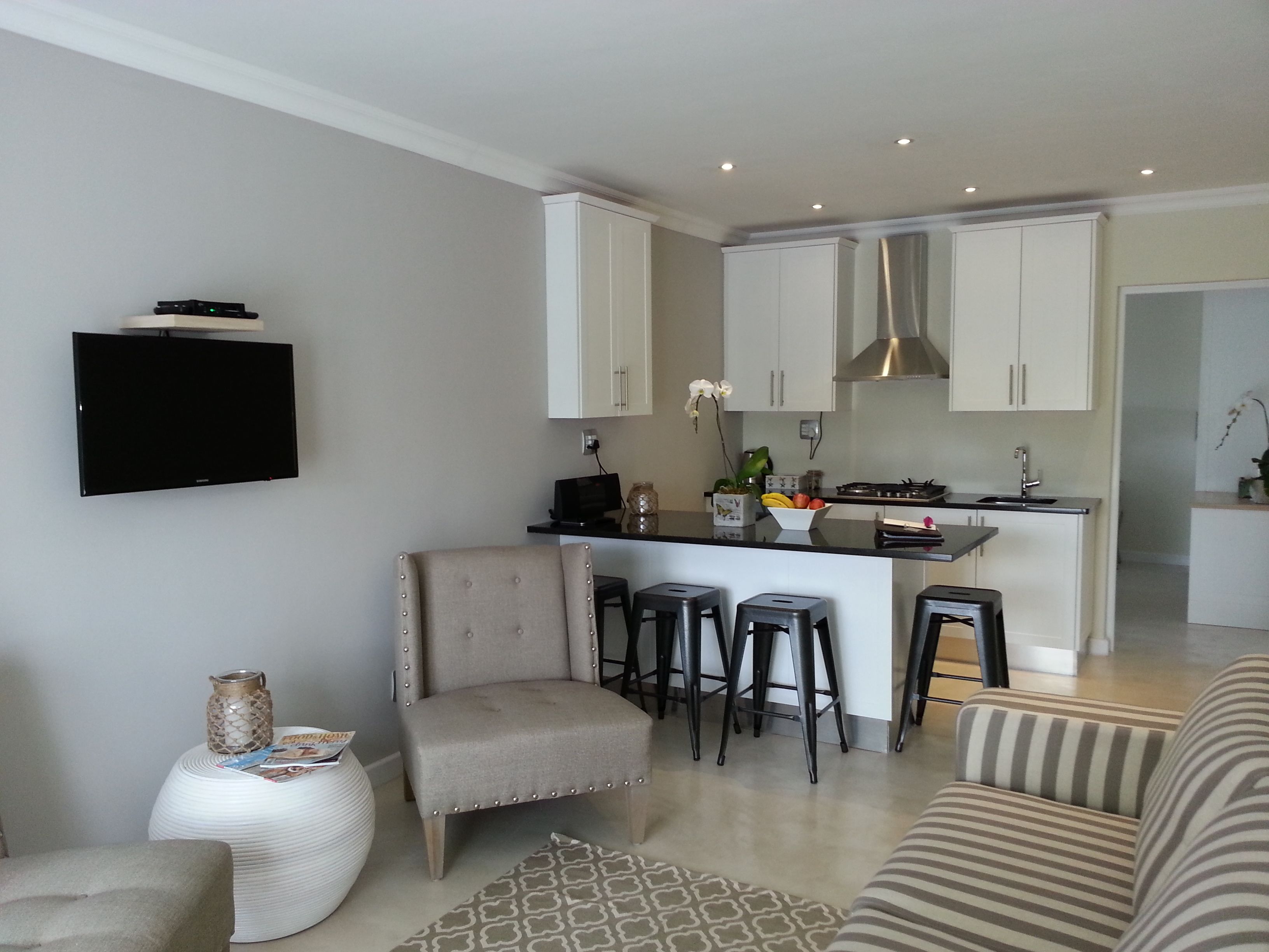 Una cuina moderna amb armaris blancs, una illa central amb tamborets, un televisor de pantalla plana al mur i una zona de menjador amb sofàs estriats.