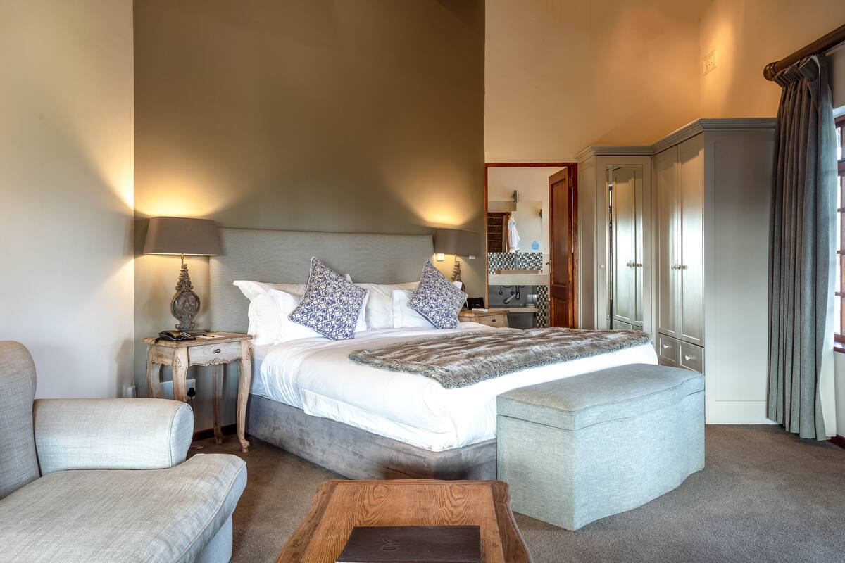 A well-lit bedroom with a large bed, a nightstand with a lamp, a wooden coffee table, and a grey ottoman.