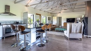 A spacious kitchen with modern appliances, a dining area with bar stools, a living room with a sofa and TV, and a large window offering a view of greenery. - Wedgeview Country House & Spa (Stellenbosch)