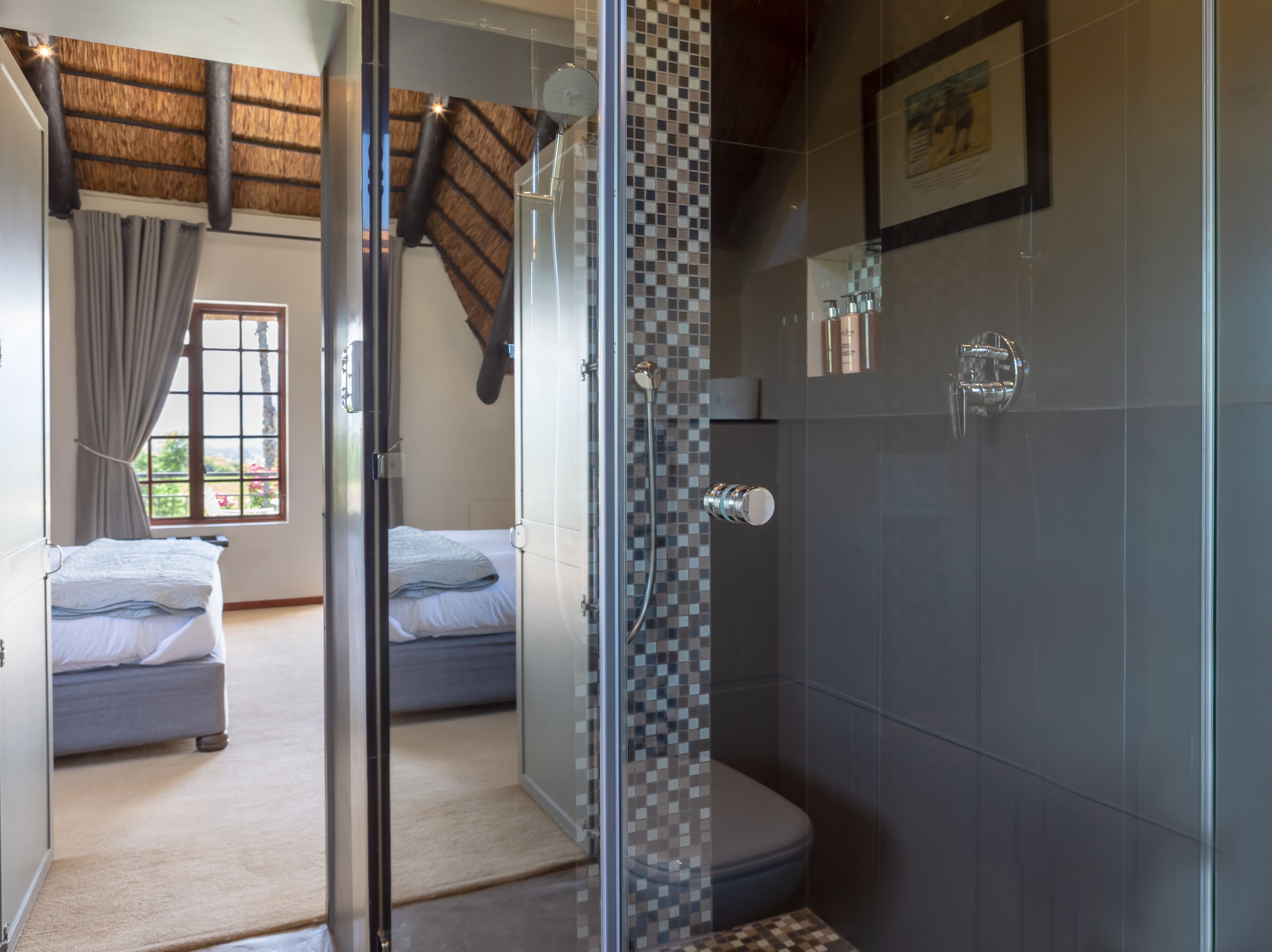 A modern bathroom with a shower, a toilet, and a glass-enclosed shower area. There is a bed with a grey headboard in the adjacent room.