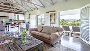 A spacious living area with a beige sofa, a rustic coffee table, and a view of the outdoors through large windows. - Wedgeview Country House & Spa (Stellenbosch)