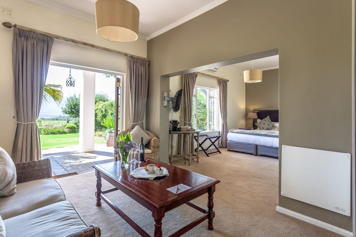 A spacious living area with a wooden coffee table, a sofa, and a dining table. There is a large window with curtains, a television mounted on the wall, and a bed in the background.