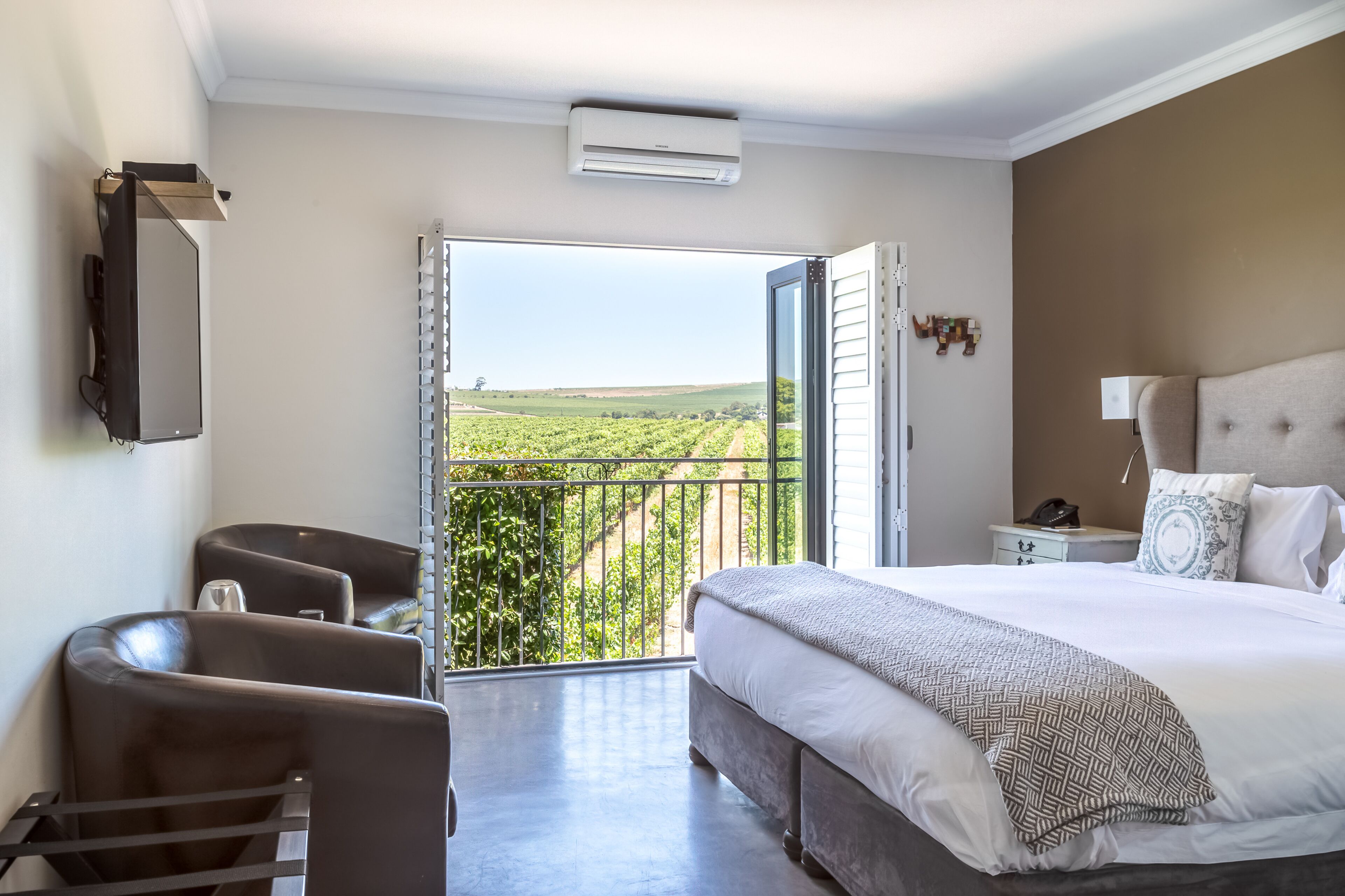 Ein Schlafzimmer mit einem großen Bett, einem Schreibtisch und einem Sessel, ein Balkon mit Blick auf ein Weinberg und ein an der Wand befestigtes Fernsehgerät.