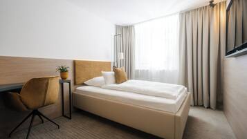 A hotel room with a bed, a desk, a chair, a potted plant, and a window with curtains.