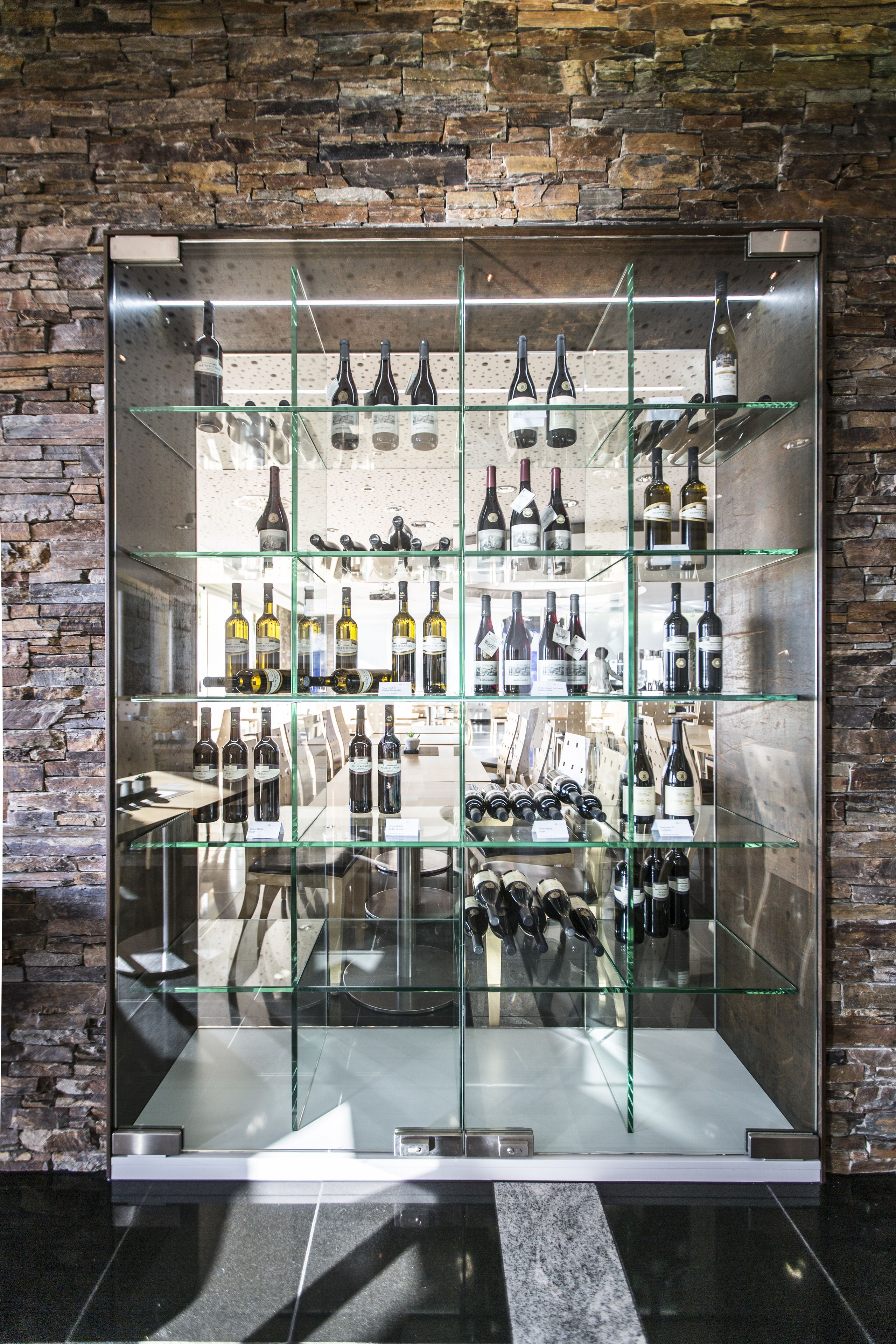 A wine cellar with multiple shelves of wine bottles, a stone wall background, and a reflective floor.