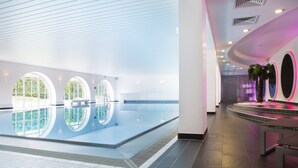 A modern indoor swimming pool with large windows, a tiled floor, and a seating area with a curved bench. - Hotel Schloss Montabaur (Montabaur)