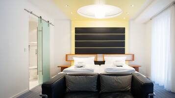 A modern hotel room with a bed, a sofa, a glass door leading to a bathroom, and a large window with curtains.