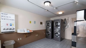 A laundry room with two stacked front-loading washing machines, a stainless steel refrigerator, and a shelf with folded towels and detergent bottles. - Knights Inn South Amboy (South Amboy)