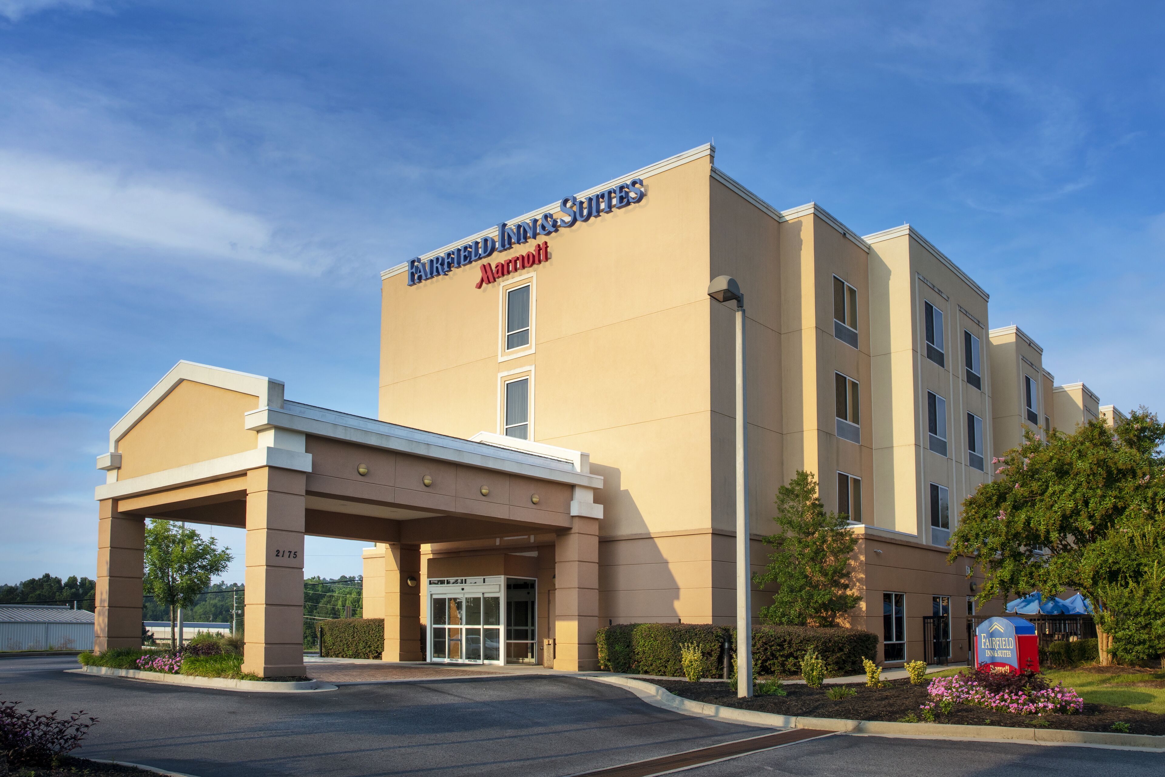 A Fairfield Inn & Suites by Marriott hotel with a beige exterior, a covered entrance, and a landscaped garden with flowers.