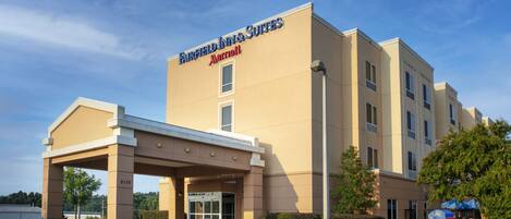 A Fairfield Inn & Suites by Marriott hotel with a beige exterior, a covered entrance, and a landscaped garden with flowers.