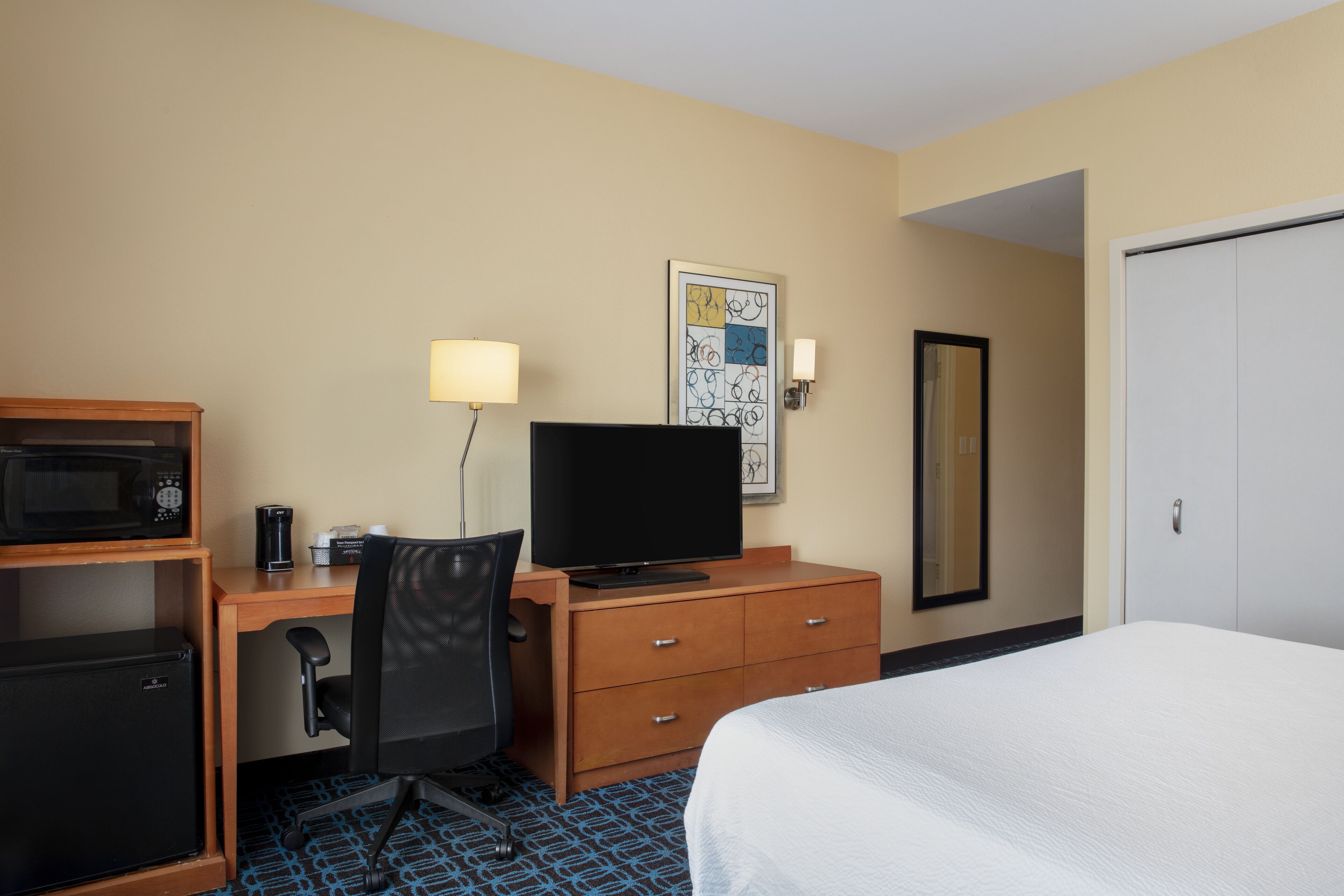 A hotel room with a bed, a desk with a television, a microwave, a refrigerator, a chair, and a wardrobe.