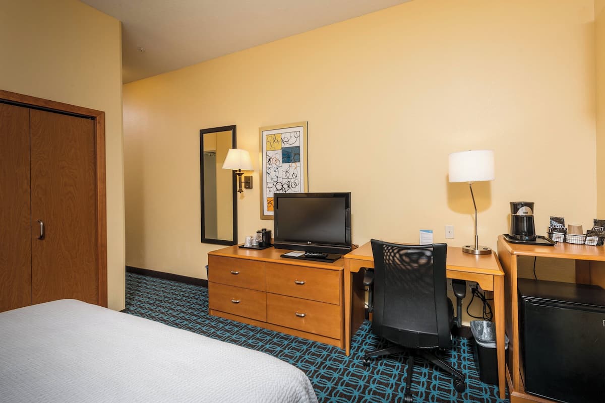 A hotel room with a bed, a desk with a computer, a chair, a lamp, and a small refrigerator.