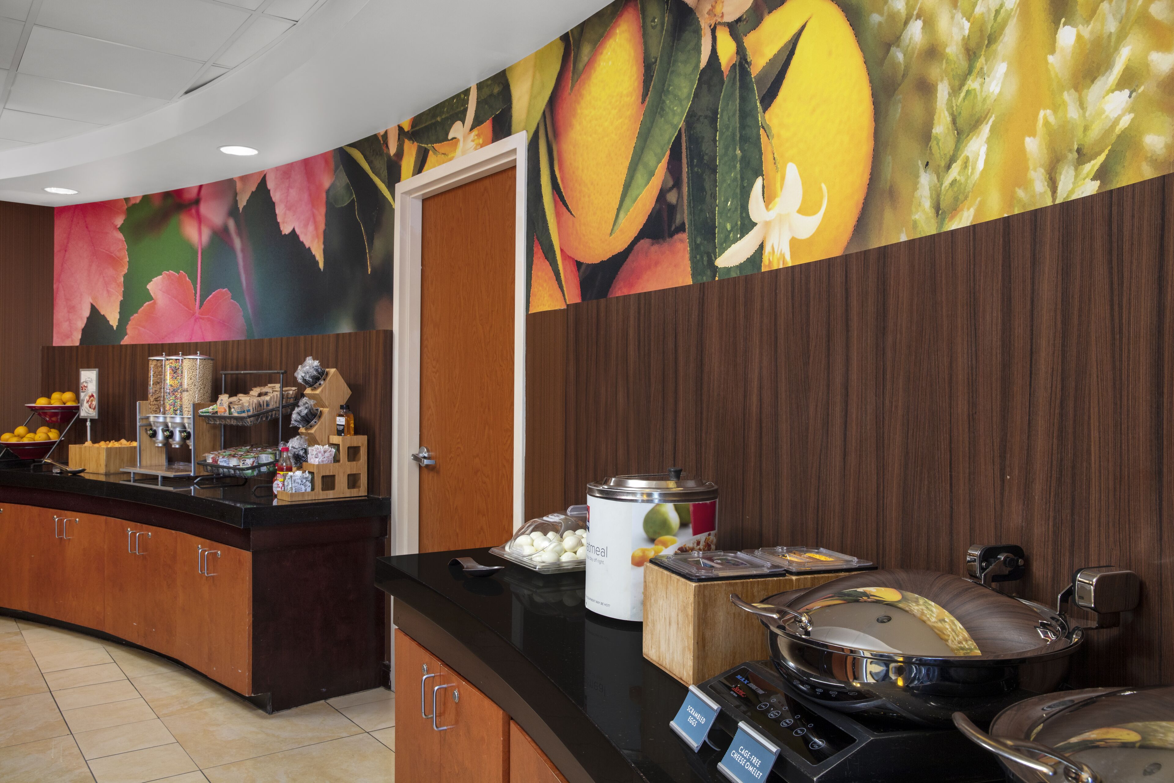 A breakfast buffet area with a variety of food items, a coffee station, and a decorative wall with fruit and floral patterns.