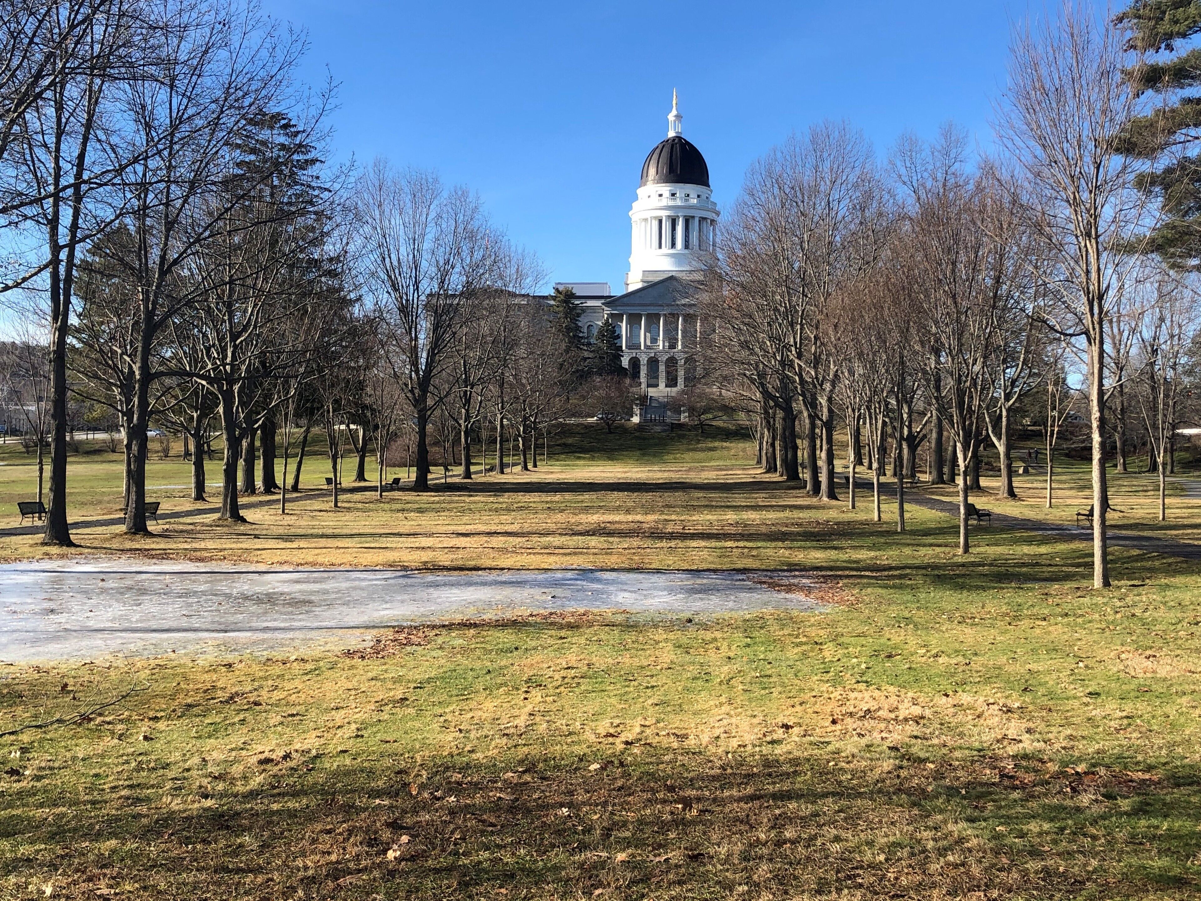 A park with a large white building featuring a prominent dome, surrounded by trees and benches.