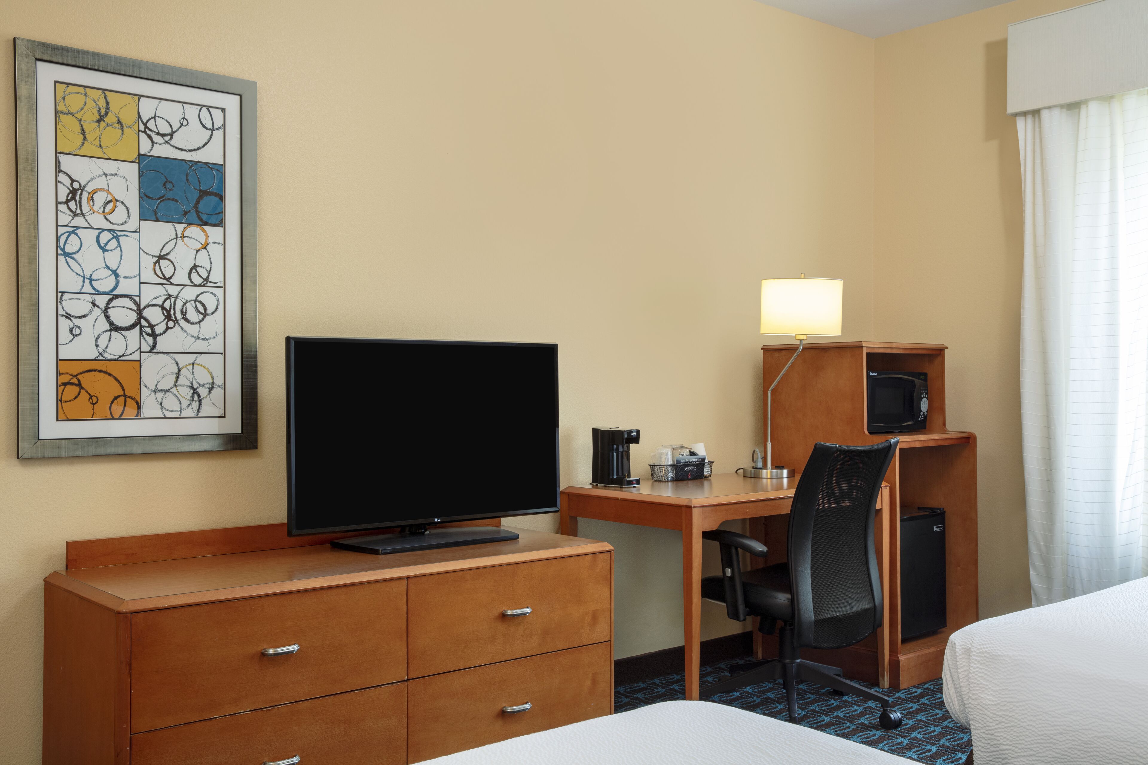 Habitación de hotel con cama, escritorio con computadora, televisor, silla, refrigerador pequeño y una obra de arte enmarcada en la pared.