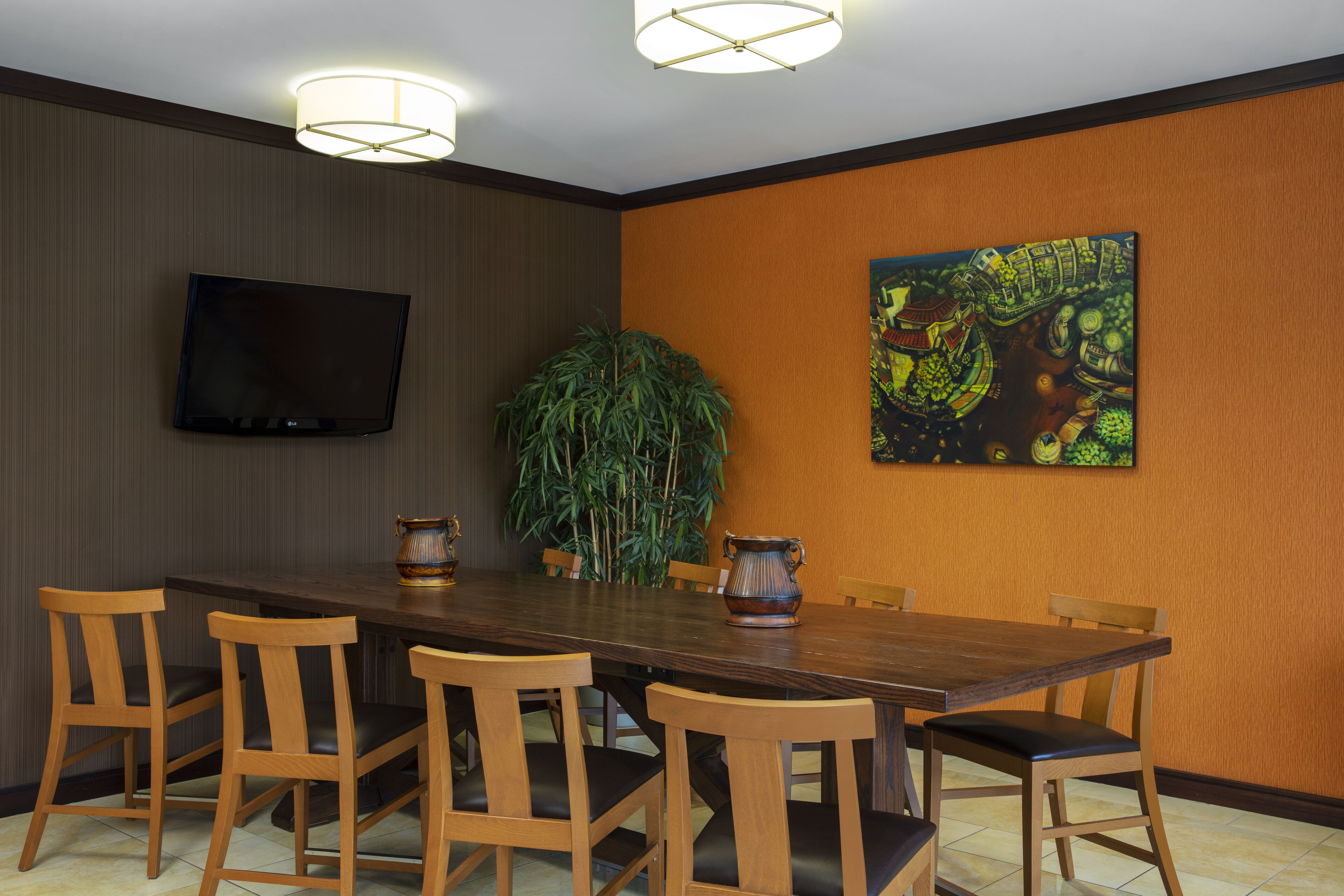 A dining area with a long wooden table, six wooden chairs, a flat-screen TV mounted on the wall, a large potted plant, and a colorful painting.
