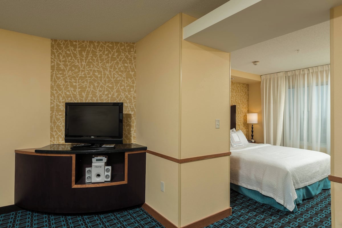 A hotel room with a flat-screen TV on a wooden console, a bed with a white comforter, and a patterned carpet.
