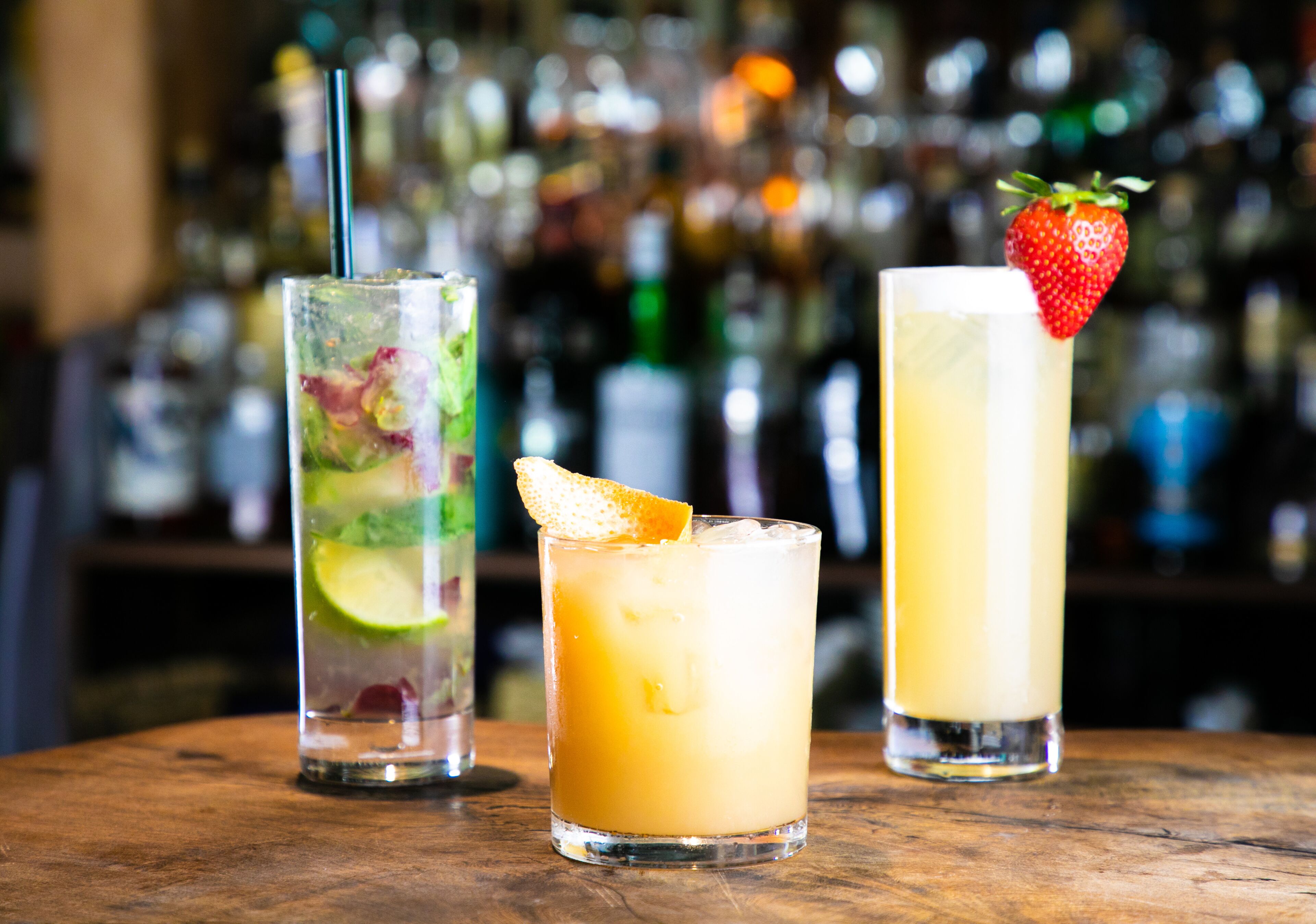 Three cocktails on a wooden bar counter with a blurred background of bottles.