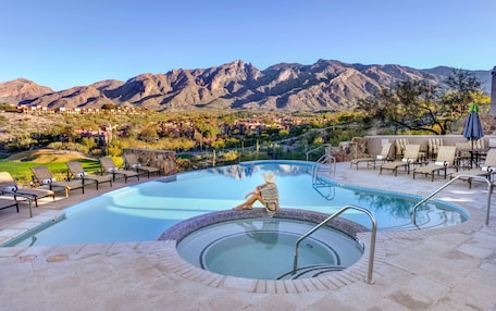A pool area with lounge chairs and a hot tub, surrounded by mountains and a view of a town.