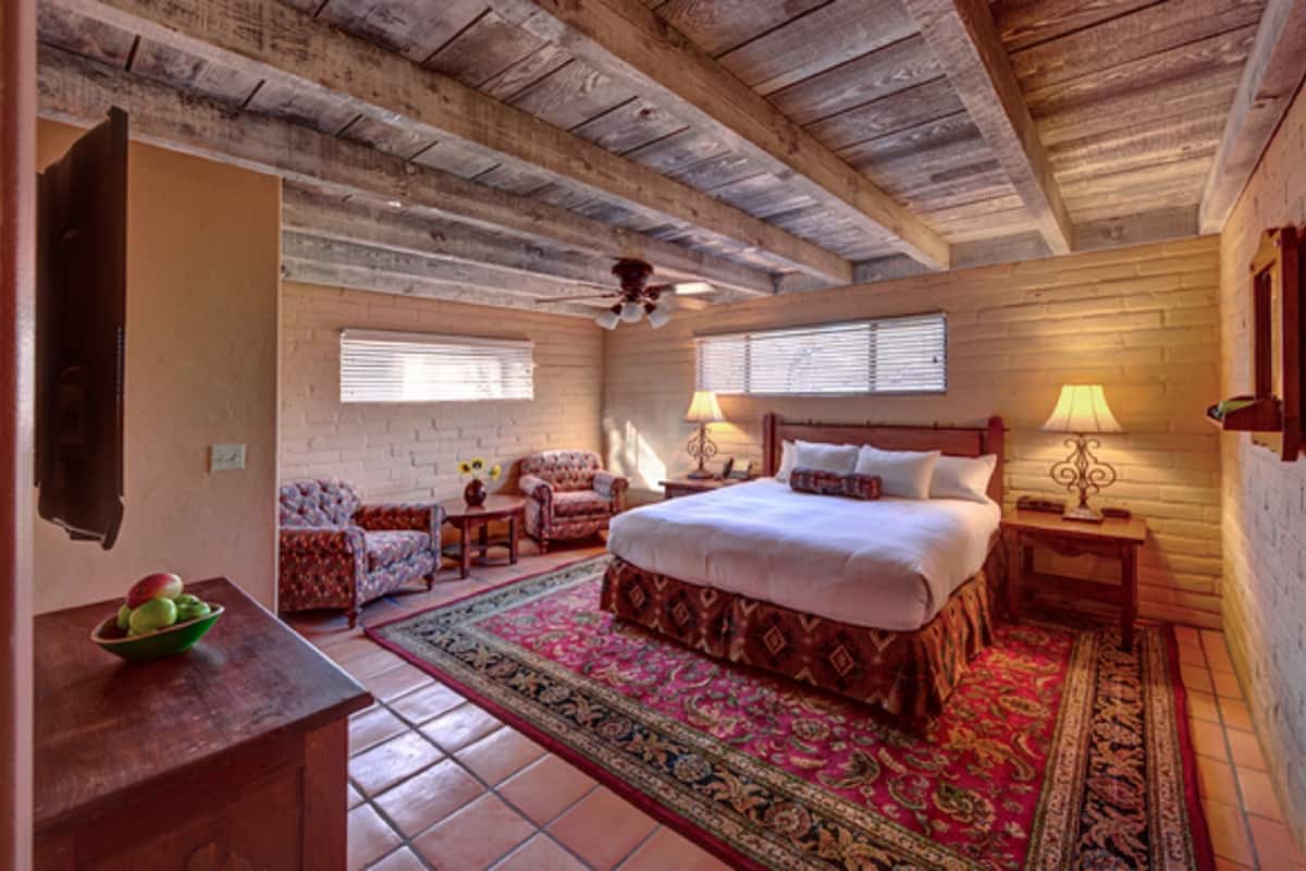 Une chambre avec un plafond en bois, un grand lit, deux tables de chevet avec des lampes, une chaise et une petite table sur laquelle se trouve un saladier rempli de fruits.